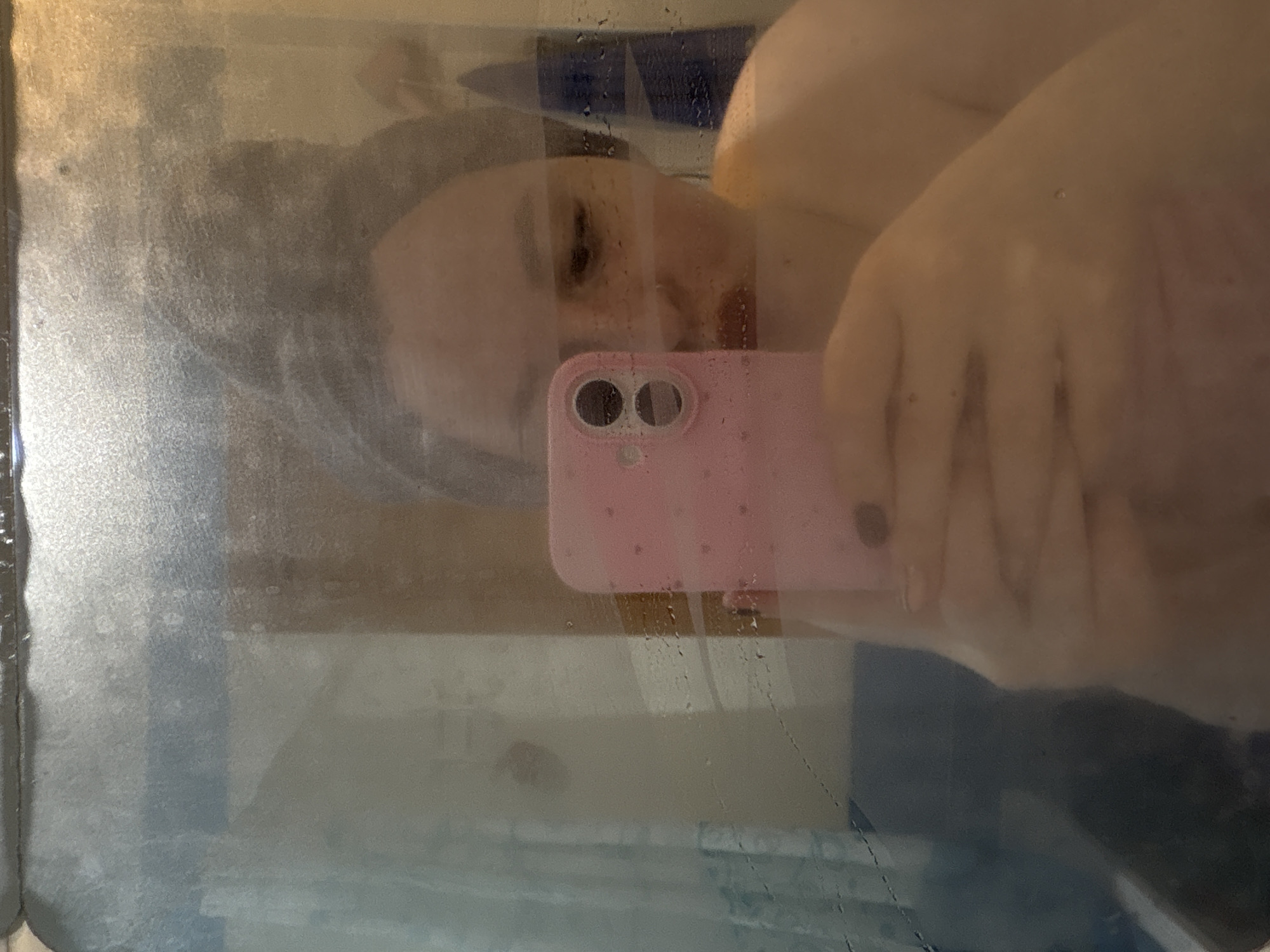 A person is taking a selfie in a shower. The person is holding a pink phone and is wearing a shower cap. The location cannot be determined based on the image alone.