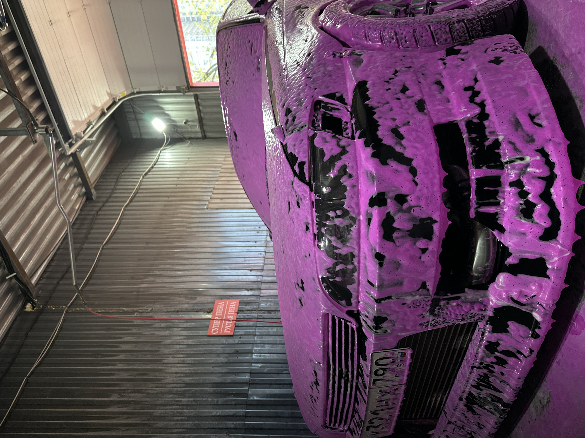 The image shows the front of a black car covered in pink foam at what appears to be a car wash. The car's license plate, which is partially obscured by foam, reads "K321HX 790 FJS". The license plate indicates that it is from Russia. A red and white sign with Cyrillic text is visible on the wall, indicating a product name or instructions related to car washing. The car wash is located in a building with corrugated metal walls. A light fixture with a visible bulb illuminates the location. Based on the license plate information and prior context, the location is Khimki, Russia.