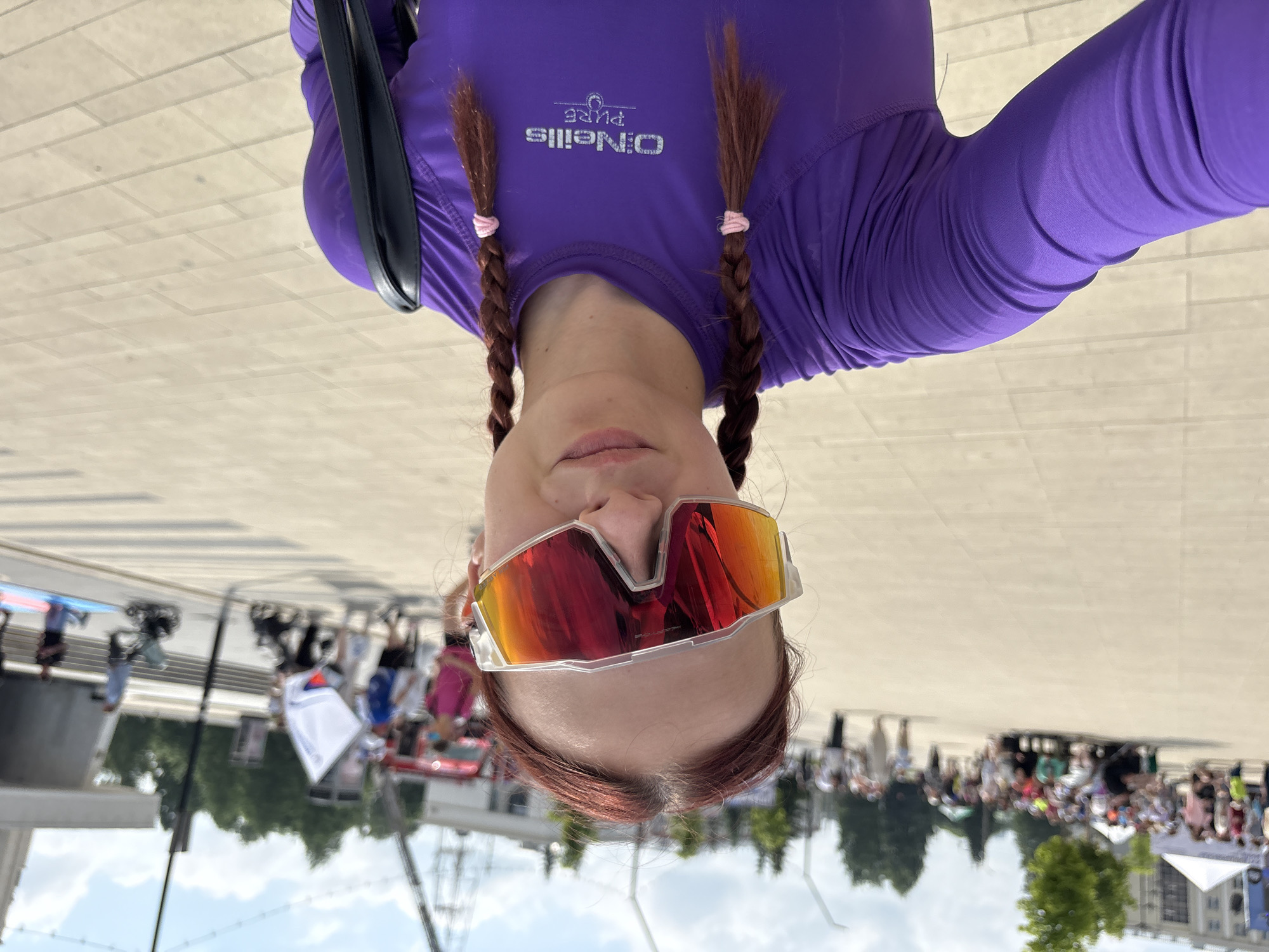 A young woman with reddish-brown hair styled in two braids, wearing a vibrant purple long-sleeved shirt with a white logo on the chest, and oversized, reflective sports sunglasses, takes a selfie outdoors on a paved plaza. In the background, a crowd of people are gathered, some seated, others standing. A white tent is visible to the left, and trees and buildings line the horizon. The sky is partly cloudy. In the mid-ground, a red fire truck and several individuals are present, including someone holding a white flag with Russian colors. The ground is paved with light-colored rectangular tiles. The overall scene suggests a public event or gathering in an urban setting. The location is identified as Izhevsk, Russia.