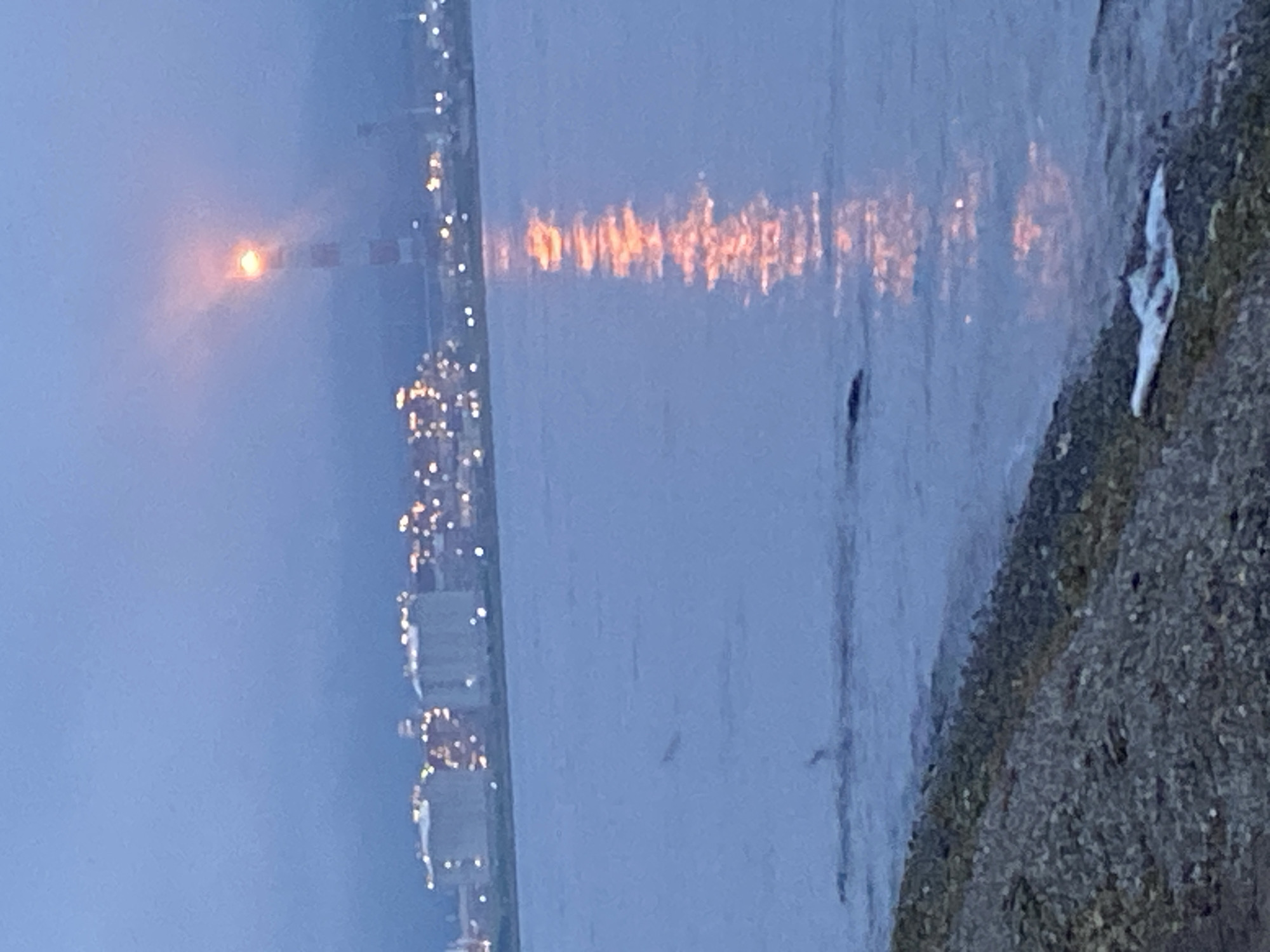 The image depicts an industrial waterfront scene at dusk or dawn in Korsakov, Russia. In the foreground, a stony embankment lines the edge of a calm body of water. The water reflects the lights of a distant industrial complex situated across the bay. These structures appear to be large storage tanks or processing facilities, illuminated by numerous small, warm lights. Dominating the skyline is a tall, red-and-white striped industrial chimney or tower, with a bright, glowing light at its apex, possibly a warning beacon or flare. The sky is a muted blue-grey, suggesting low light conditions. In the water, a dark shape, potentially a buoy or debris, is visible.