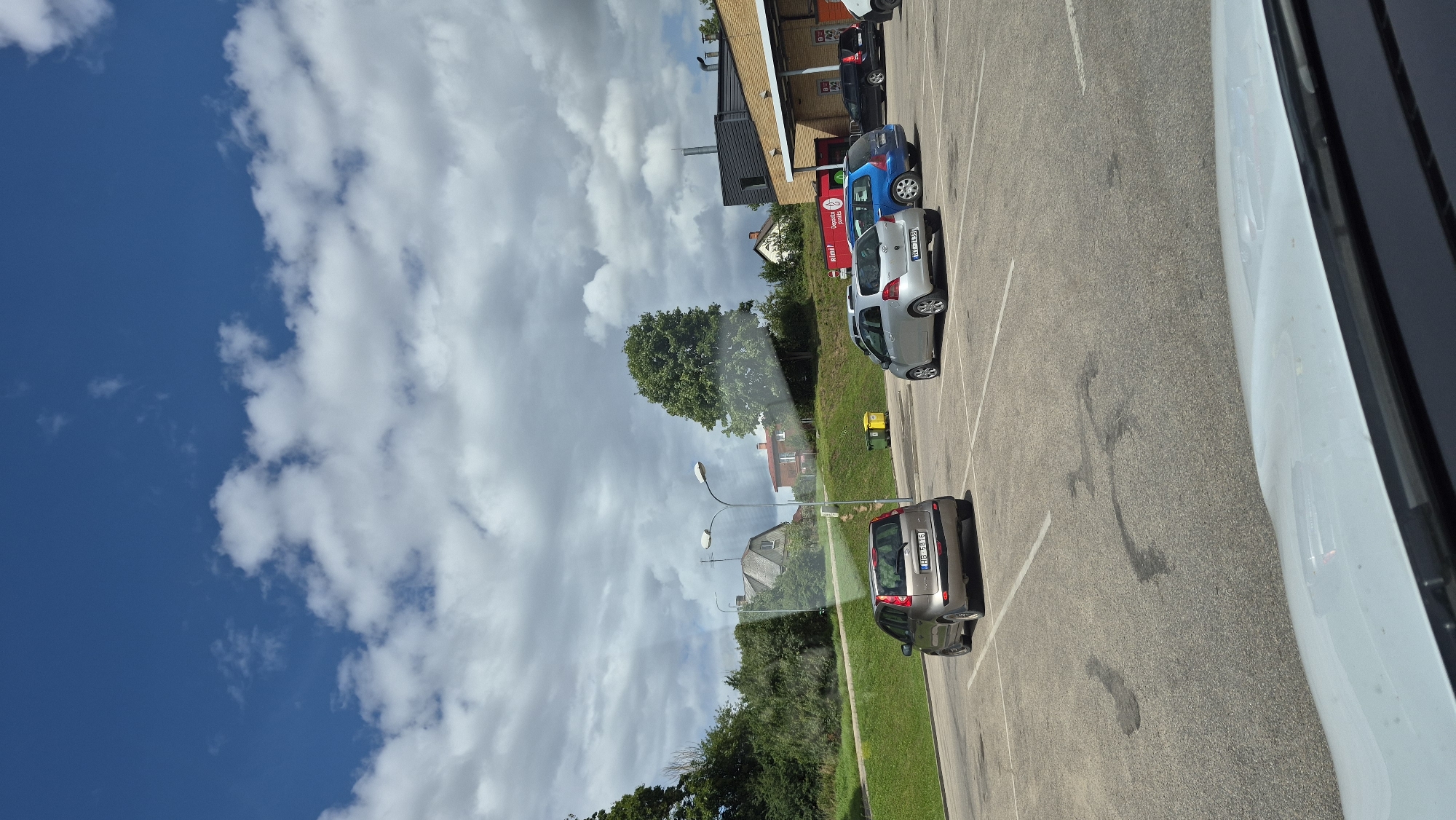 This is a photograph taken from inside a vehicle, looking out at a parking lot. The parking lot is paved with asphalt and marked with white lines indicating parking spaces. Several cars are parked in the lot, including a silver hatchback in the foreground, a silver hatchback parked further back, a blue hatchback, and a black SUV. In the background, there is a building with a yellow facade and a dark roof, which appears to be a commercial establishment. Behind the building, a green hill slopes upwards, covered with trees. A few houses are visible among the trees. Two streetlights stand in the middle of the parking lot. The sky above is blue with scattered white clouds. The image quality is slightly distorted due to reflections on the vehicle's windshield. Based on the visible license plates (HB 5816 and NT 303B) and the context, this scene is likely located in Jēkabpils, Latvia.
