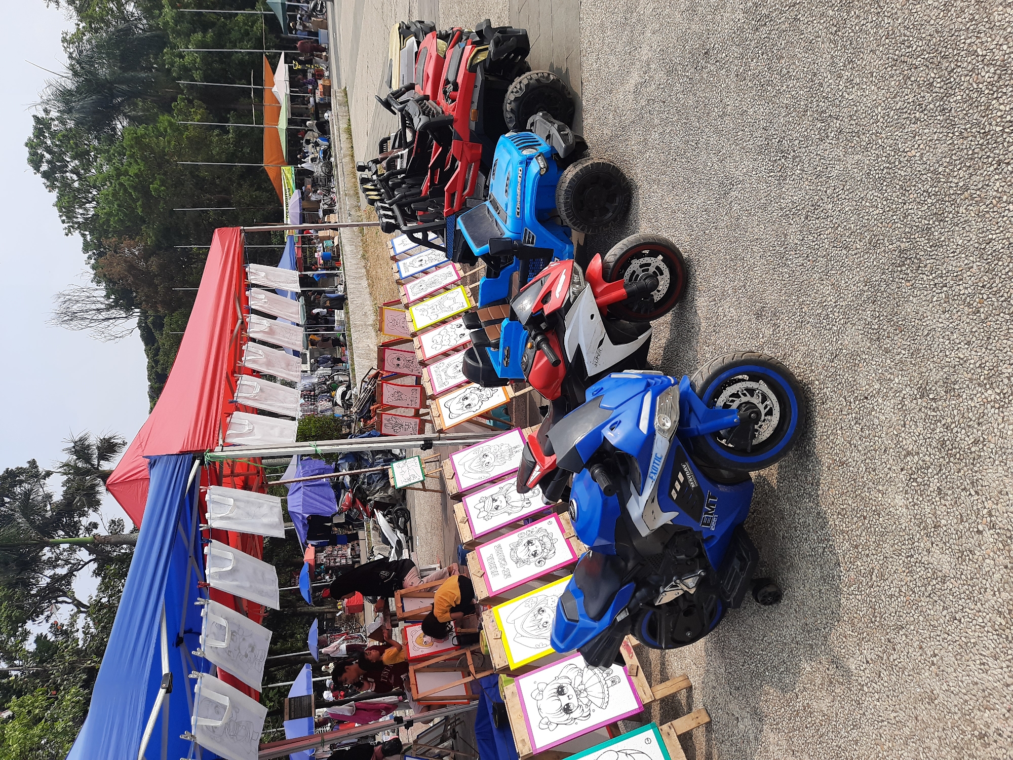 This image depicts an outdoor marketplace event, likely a weekend fair or festival, set up in a park-like environment. The scene is dominated by a row of colorful canopies, red and blue in the foreground, stretching back to other stalls with orange, green, and white awnings.
In the immediate foreground, several children's ride-on vehicles are displayed, including a bright blue electric motorcycle with the brand "EXOTIC" visible, and a blue electric jeep labeled "COMMANDO". Behind these are a red and white electric motorcycle and several red and black electric ATVs.
In front of the canopies, a row of easels holds coloring pages featuring cartoon characters, suggesting an art activity for children. Several children and adults are present, with some children actively engaged in coloring or observing the drawings. A woman in a black hijab is seen supervising at one of the easels. The ground is covered in a textured, gravel-like surface.
In the background, trees form a dense backdrop, indicating a location with significant greenery. Beyond the canopies, more stalls and activity can be discerned, with people browsing and interacting. The overall atmosphere appears lively and family-oriented. The presence of various food and merchandise stalls, combined with recreational activities for children, is characteristic of public gatherings and markets often held in parks within urban areas.
While the exact location is not explicitly stated, the combination of the market setup, the presence of trees, and the general ambiance aligns with common public spaces in cities like Bandung, Indonesia, which are known for hosting such weekend events in their parks and recreational areas.