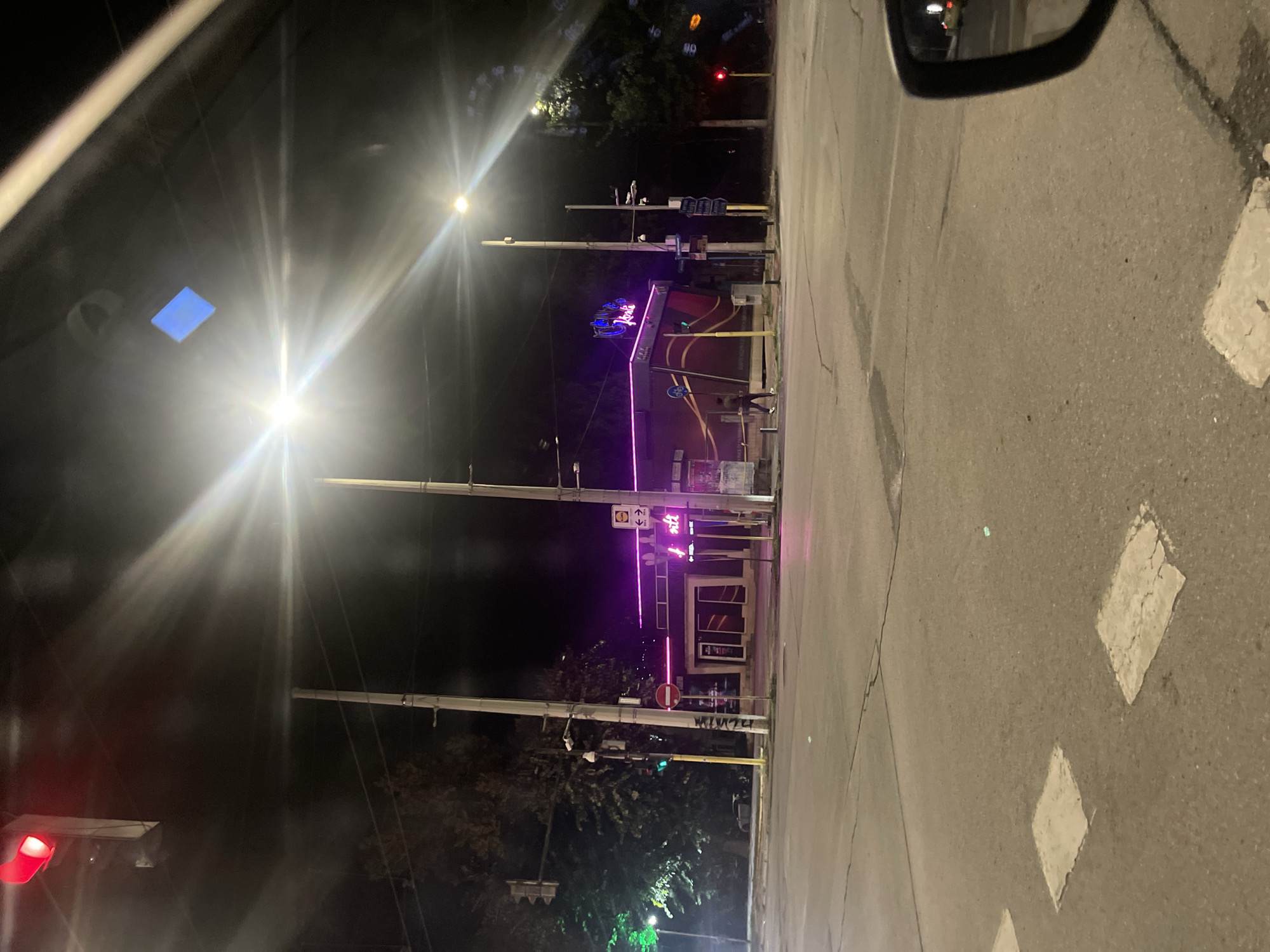 The image shows a street intersection at night, likely in Ruse, Bulgaria, given the visible signage. A building with vibrant purple neon lights outlining its facade and spelling out "KOKO" is prominent in the center. A person is walking on the sidewalk in front of this building. Streetlights and traffic signals are visible, with one red traffic light illuminated. The road surface shows painted white lines and some patching. The scene is captured from a low-angle perspective, possibly from inside a vehicle, as a car's side mirror is visible in the bottom right corner. The overall atmosphere is nocturnal, with artificial lighting dominating the scene.