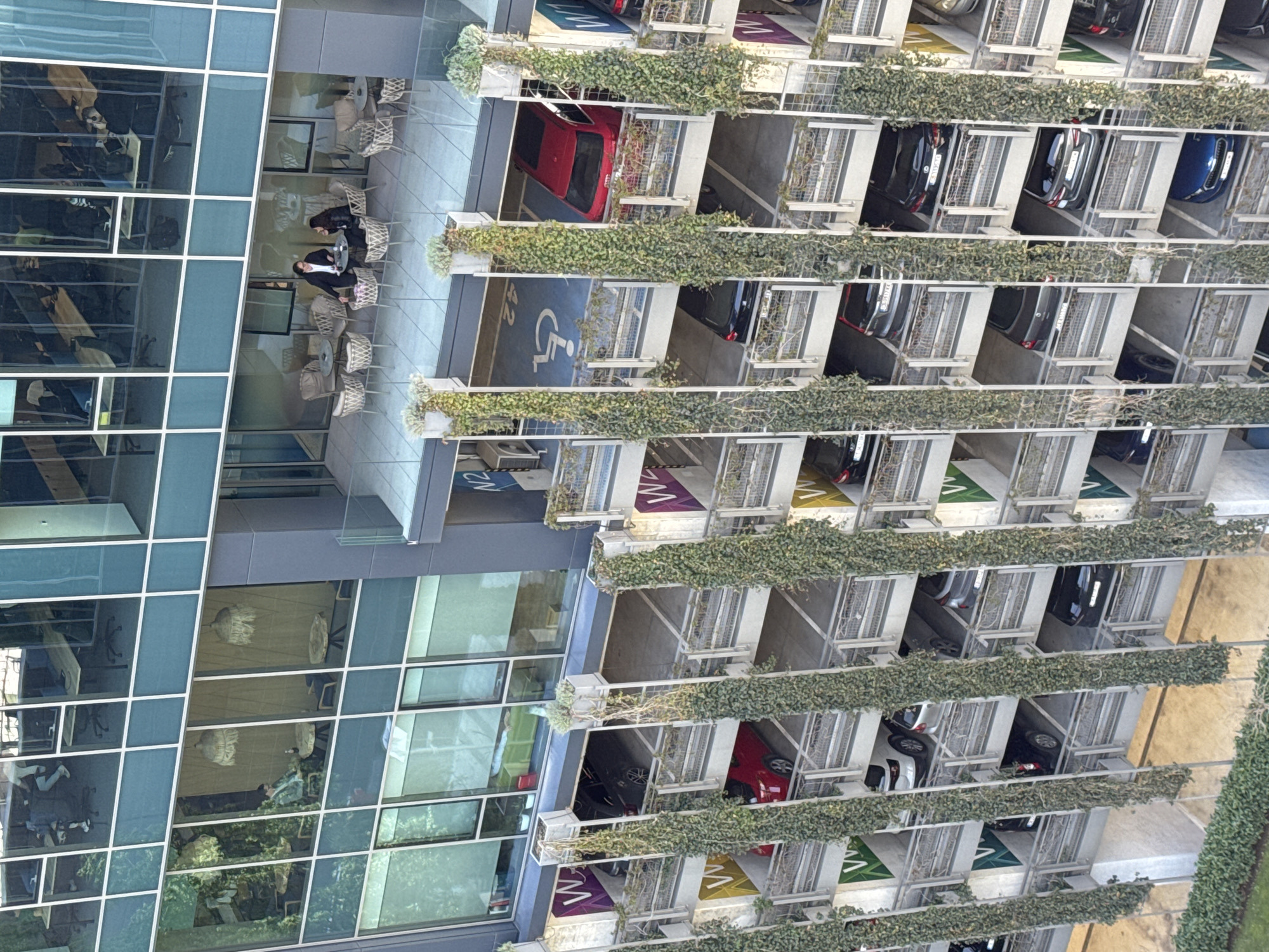 A daytime aerial or high-angle shot captures a modern multi-story building complex in Sofia, Bulgaria, featuring a glass-paneled office building, an outdoor terrace, and an integrated multi-level parking garage. The lighting suggests clear, sunny weather.
On the left, the contemporary office building exhibits a grid of reflective glass windows, showcasing reflections of indoor office environments with desks and chairs. In the middle ground, an outdoor terrace, possibly belonging to a café or shared office space, is furnished with several round tables and rattan-style chairs. Two people are seated at one table, appearing to be engaged in conversation or a meeting. One wears a dark jacket, the other a white top.
To the right, a multi-story parking garage extends vertically, with cars parked on various levels. The garage's structure is adorned with lush green climbing plants, possibly ivy, along its edges, adding a natural element to the concrete architecture. Visible cars include red, black, blue, and white models.
Notable visible text includes large letters painted on the parking levels, such as "W" and "V", likely indicating sections or floor numbers. One level clearly shows a blue handicapped parking space symbol with a wheelchair icon and the number "42". On an upper section of the building's facade, "N2" is also visible. The scene depicts a typical busy daytime environment in a modern urban setting.