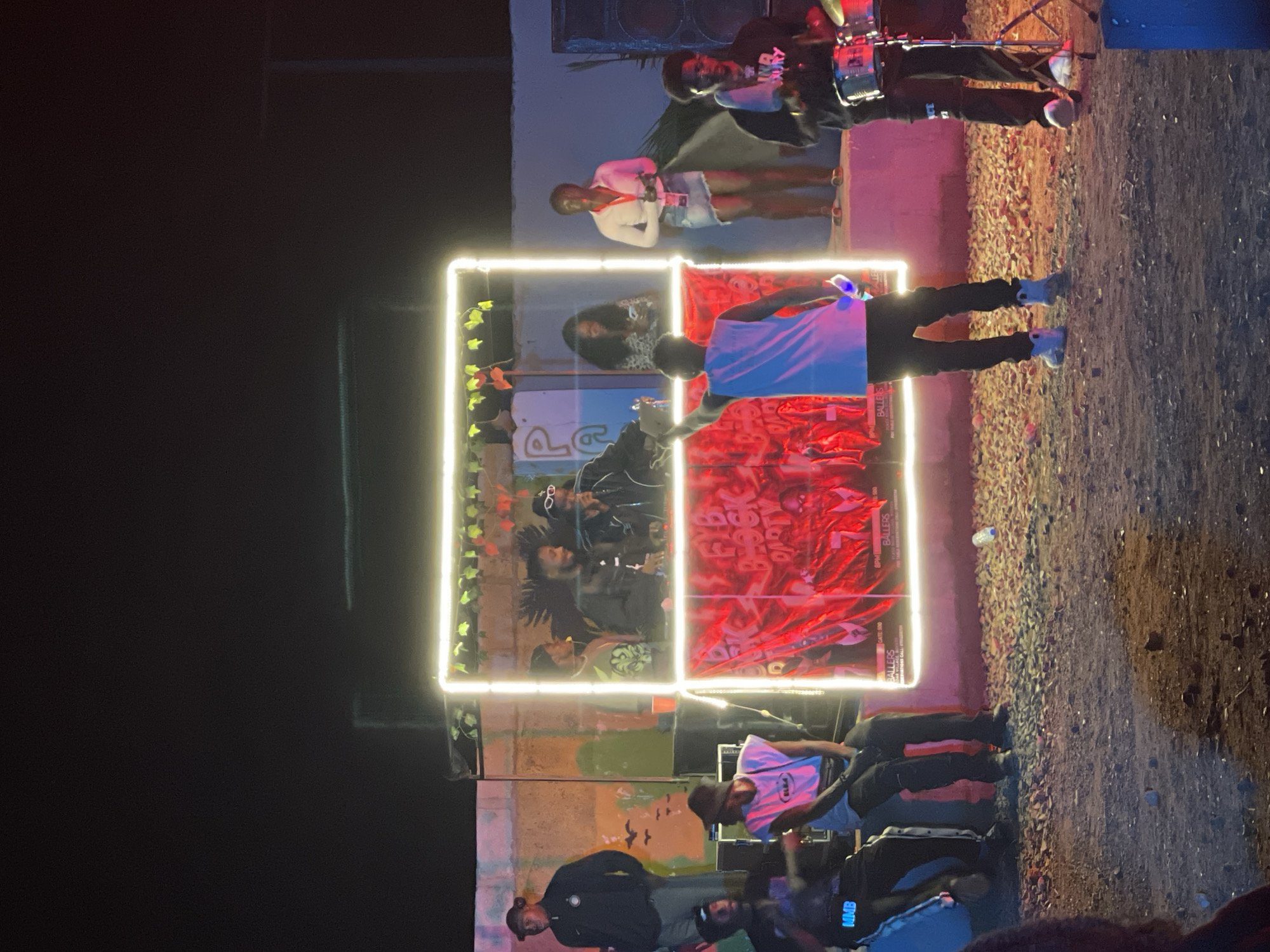 The image depicts an outdoor event, likely a block party, at night in Durumi, Nigeria. A brightly lit stage area, outlined in neon white light, serves as the focal point. On the stage, several individuals are present. Two men are visible behind a counter, one appears to be speaking into a microphone, while the other is looking at him. Another man is standing to the left of the stage, facing forward. A woman in a white shirt and denim skirt stands on the right side of the stage, holding a phone. In front of the stage, several people are gathered. A man in a white tank top and black pants is facing away from the camera, with his arm raised, holding a glowing object, possibly a phone or light. To his left, two men are seated on the ground, one with a microphone and the other holding a drumstick. On the far right, a man is playing drums. The ground is covered in gravel and dirt. The background consists of a painted wall and structures, suggesting an urban or semi-urban setting. The overall atmosphere appears festive and energetic.