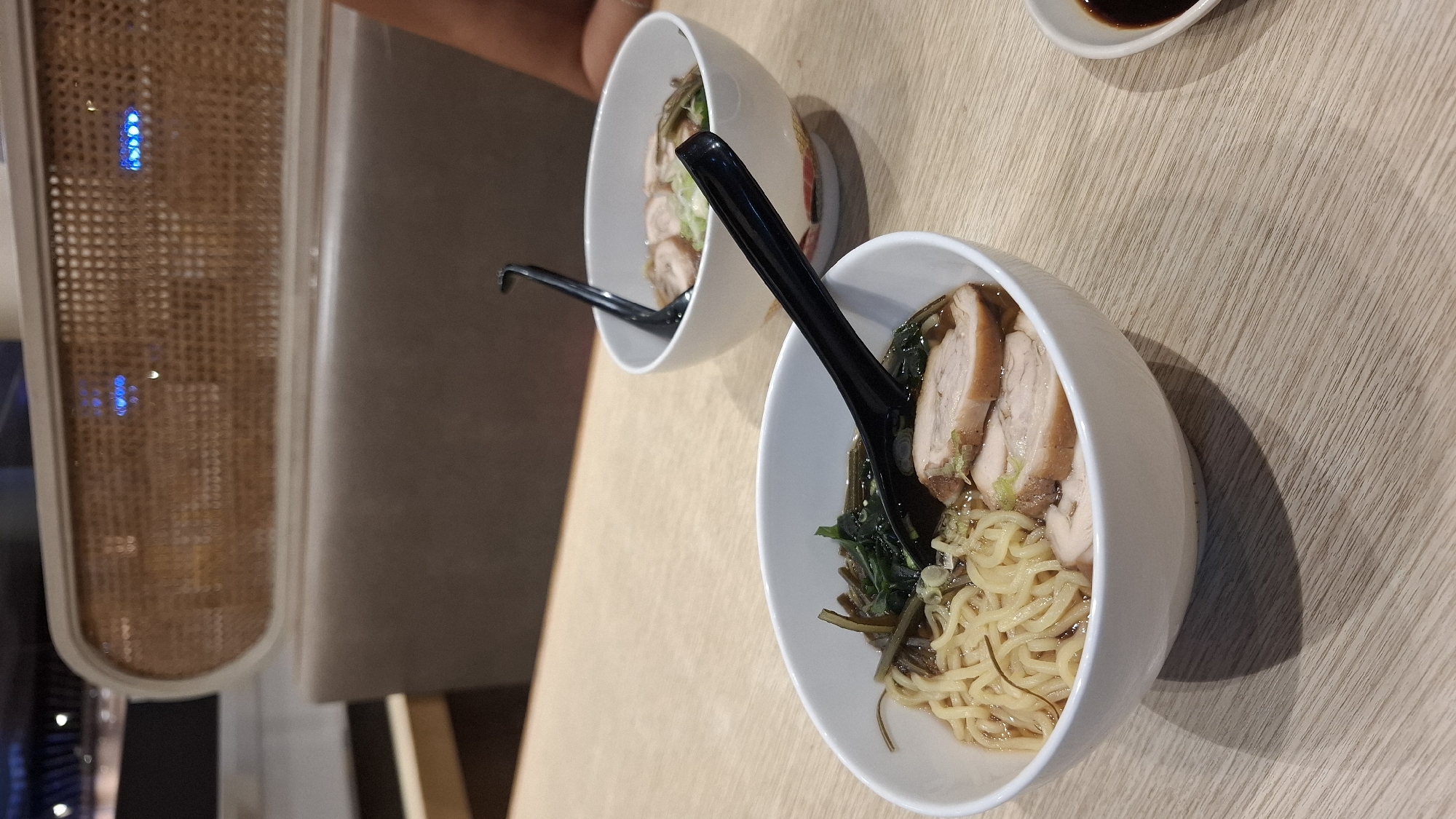 Two white bowls of ramen sit on a light-colored wooden table. Each bowl contains noodles, sliced meat, and seaweed. Black spoons are placed in each bowl. The location is potentially in Kuta, Indonesia, based on unspecified external information. A portion of a woven-patterned wall is visible in the background.