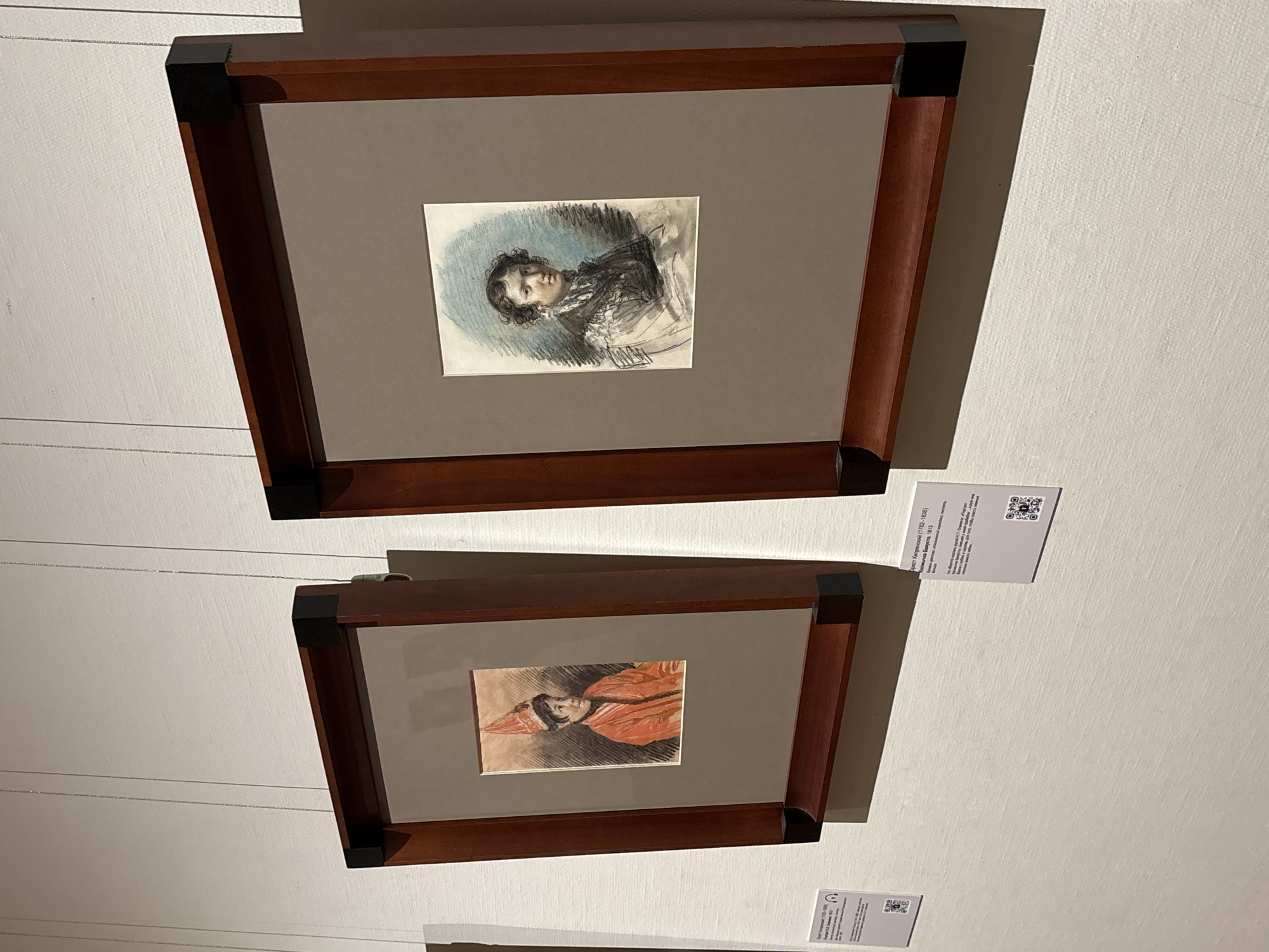 Two framed artworks are displayed on a light-colored wall. The frames are dark wood with black corners and surround what appear to be sketches or small paintings. The top artwork depicts a person with dark curly hair, wearing a dark garment with frilly white lace at the neck, sketched with blue and dark lines. The bottom artwork shows a person wearing a red hat or head covering, also done in sketch style. Each artwork has a small white information card with text and a QR code next to it on the wall. Thin, horizontal hanging wires are visible on the left side of the image, suggesting the artworks are suspended from the ceiling. Based on the information provided on the cards, it appears the artworks may be located in Moscow, Russia, although the exact location is not specified.