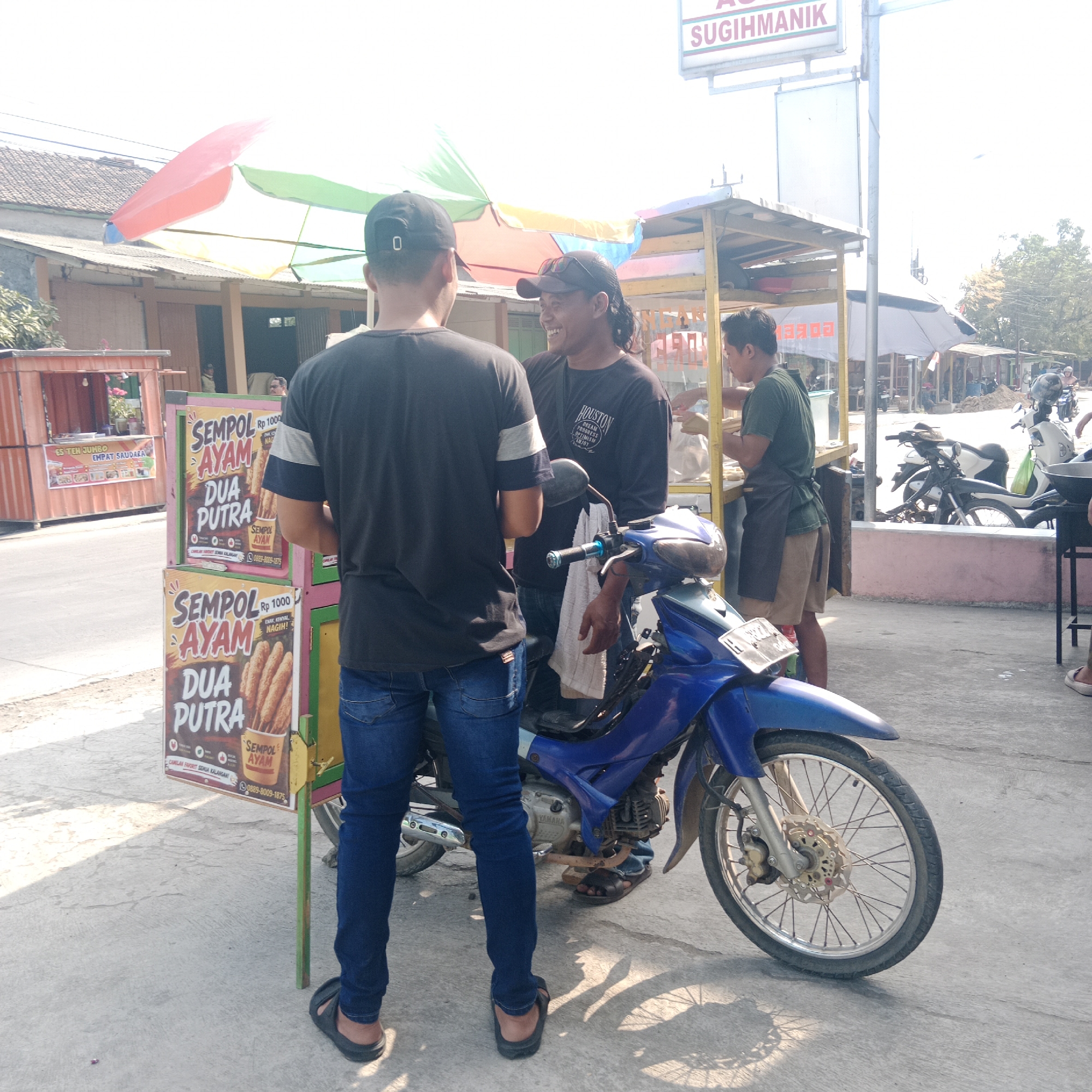 The image captures a street scene in Tegalrejo, Indonesia, with a focus on a food stall and its customers. In the foreground, two men are engaged in conversation near a blue motorcycle. One man, wearing a black t-shirt and blue jeans, is facing away from the camera. The other man, also in a black t-shirt and jeans, is facing the first man and smiling. A blue motorcycle is parked between them, with a white cloth draped over its handlebars.
Behind them, a food stall with colorful umbrellas is visible. Several signs on the stall advertise "Sempal Ayam Dua Putra" (Chicken Sempal Two Sons), indicating the food being sold. Another smaller stall, orange in color, is seen further down the street, with a sign that says "ES TEN JUMBO EMPAT SAUDARA" (Jumbo Ice Four Siblings).
In the background, other individuals are seen near a food cart, and parked motorcycles suggest a busy marketplace or roadside gathering. The environment appears to be an outdoor urban setting, with buildings and trees lining the street. The overall lighting suggests a bright, sunny day.