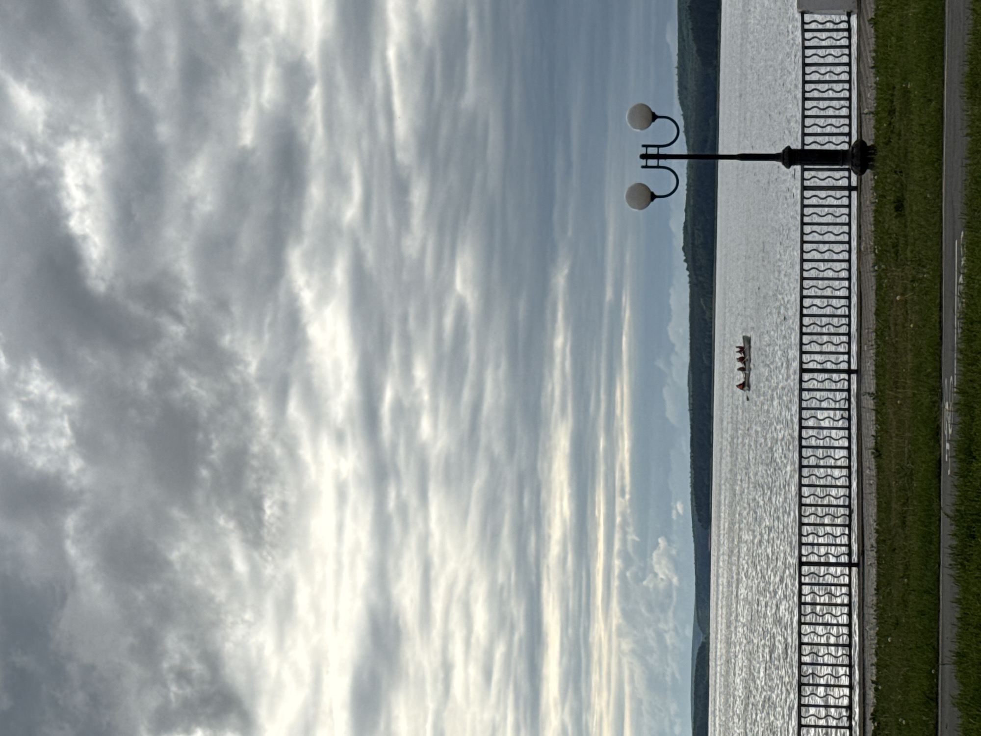 The image depicts a waterside scene in Votkinsk, Russia. A prominent cloudy sky fills the left portion of the view. To the right, a body of water stretches towards a tree-lined shore. A metal railing runs along the water's edge, with a vintage lamppost with two globe-shaped lights standing near the railing. A small boat or rowboat is visible in the distance on the water. A strip of green grass and a paved walkway are on the right edge of the image. The photo is rotated 90 degrees counter-clockwise.