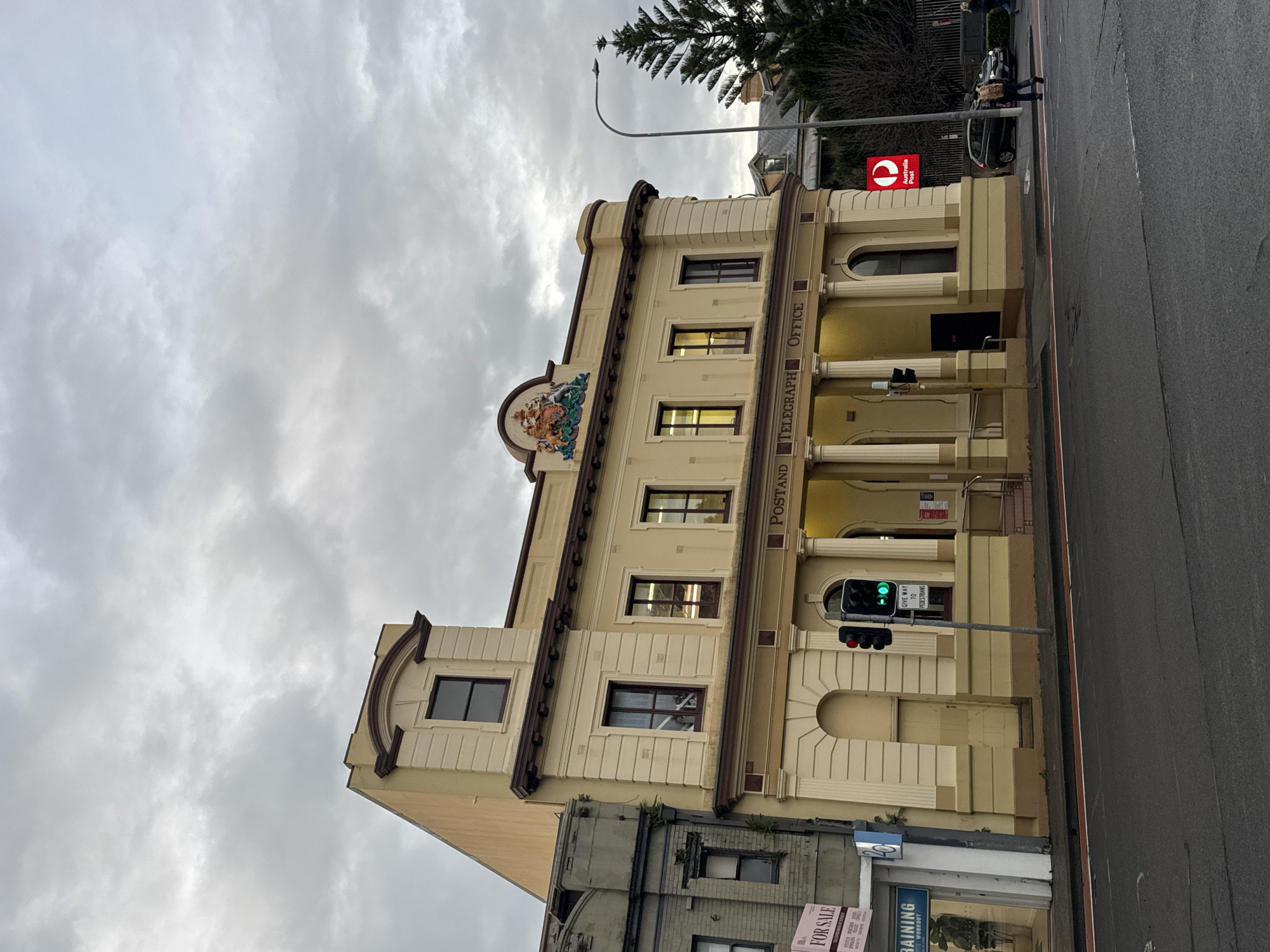 The image shows the exterior of a historic post office building, likely in Sydney, Australia, given the presence of an Australia Post logo. The building is cream-colored with brown trim and classical architectural details, including columns and arched windows. Above the entrance, a decorative crest is visible. The building is situated on a city street with a road in the foreground. A traffic light is visible, currently displaying a green signal. Several people are walking on the sidewalk and crossing the street. A car is parked on the right side of the street. The sky is overcast, suggesting a cloudy day. The left side of the image shows a neighboring building that appears older and more utilitarian, with a "FOR SALE" sign on its exterior. A blue sign indicating "TRAINING" is also visible on this neighboring structure. The Australia Post logo, a red circle with a white stylized 'P', is prominently displayed on a red illuminated sign attached to the post office building.