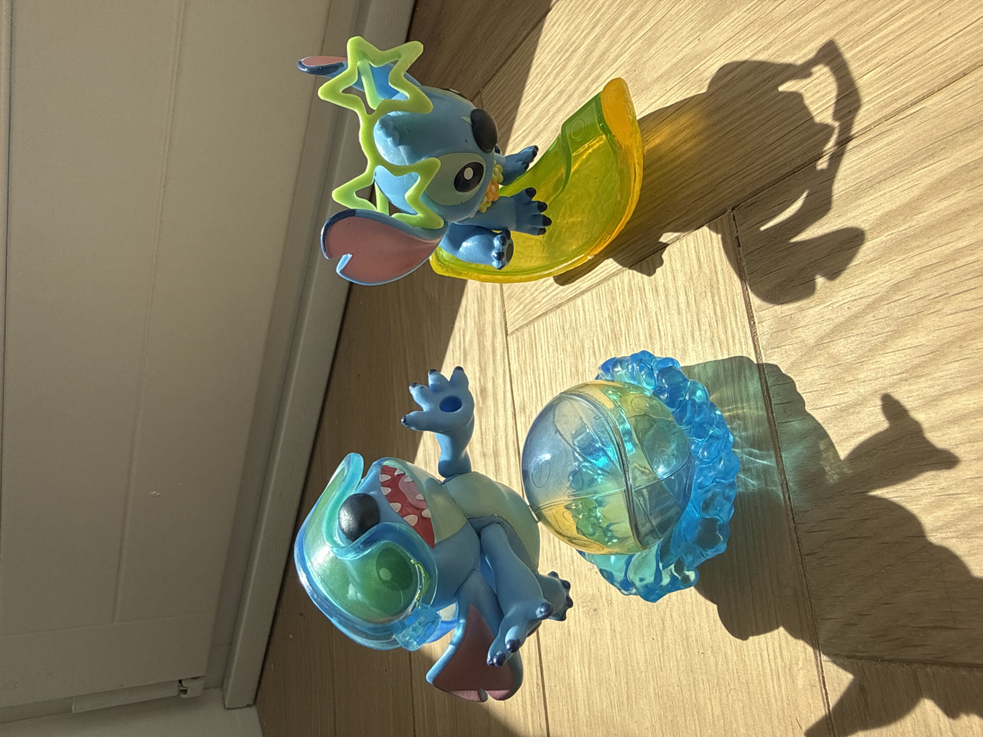 The image shows two Stitch figurines positioned on a wooden surface, likely a floor. The figurine at the top appears to be Stitch riding a yellow crescent shape with neon green stars, while the lower one seems to be emerging from a translucent blue and green orb. Both figurines cast distinct shadows on the wooden surface, indicating a strong light source. A portion of a white door or wall is visible in the upper-left corner of the image. The location and city of Moscow, Russia, cannot be determined from the provided image.