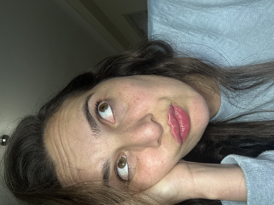A woman with brown hair and hazel eyes is captured in a close-up, tilted-angle shot. She rests her head on her hand, gazing upward. The woman's skin has freckles, and her lips are coated in a glossy, pink lipstick. She is wearing a light blue, long-sleeved shirt. The background is partially visible, displaying a white wall and a light fixture.
