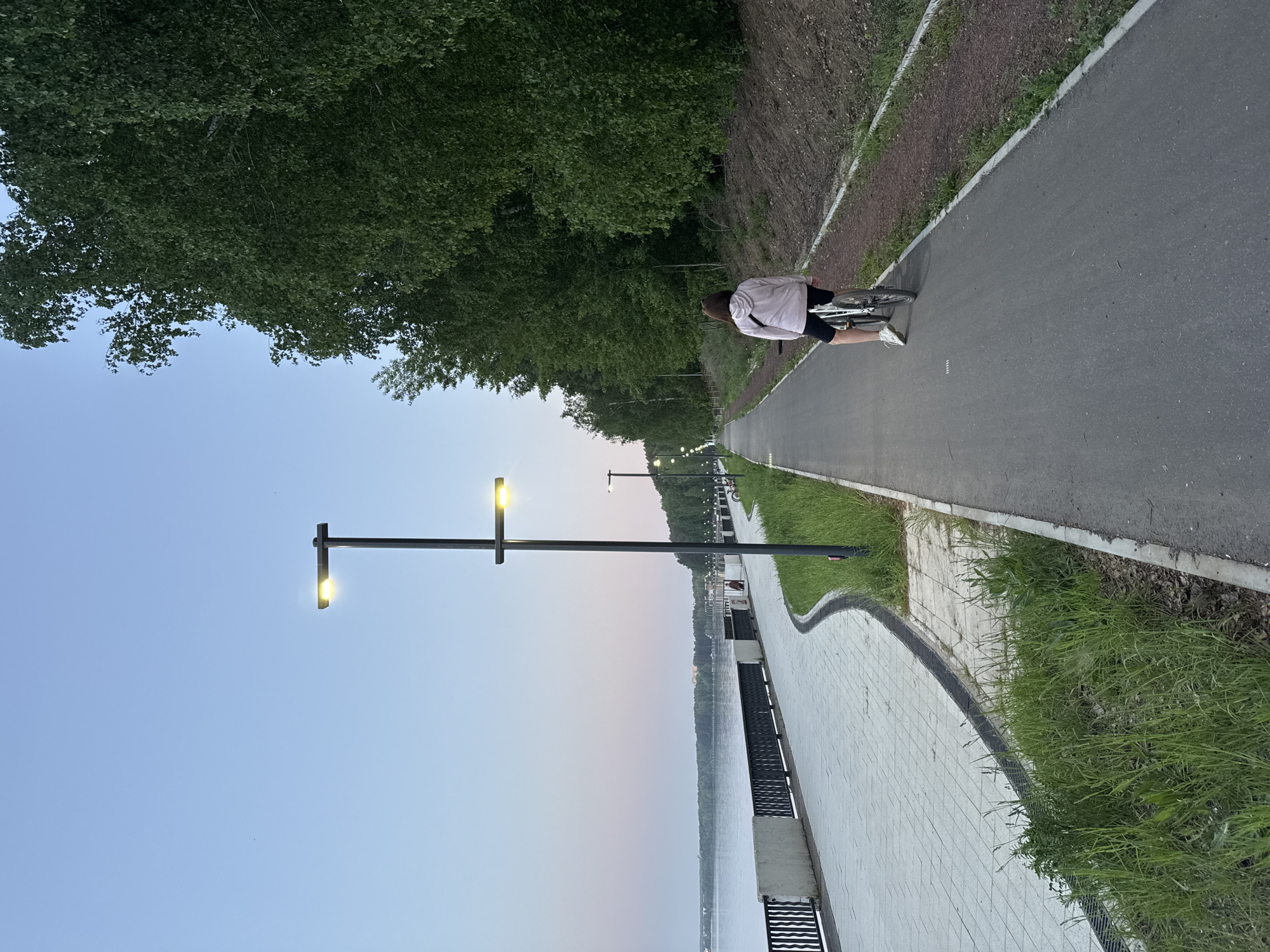 A cyclist is riding a bicycle along a paved path adjacent to a river. A row of street lights illuminate the path. The scene depicts a recreational area in Chaikovsky, Russia.