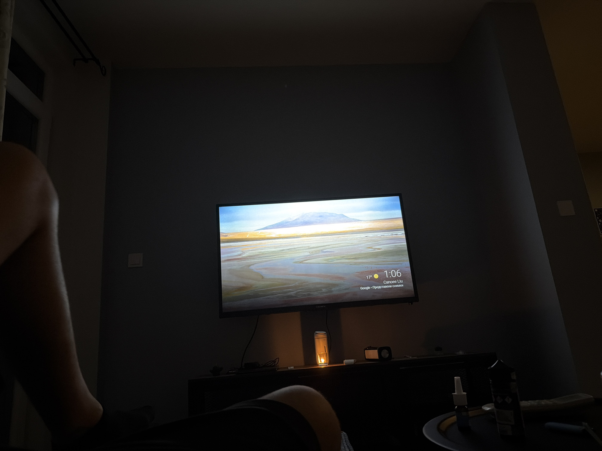 A person's leg is visible in the foreground, positioned to the left side of the frame. The leg is dark and appears to be resting. In the mid-ground, a television is mounted on a wall, displaying a landscape image of a mountain range and a body of water, with weather and time information overlaid. Below the television, a dark entertainment stand holds several objects, including a lit candle in a glass holder, a small speaker, and a bottle. The room is dimly lit, with light emanating from the television and the candle. The walls are painted a dark color. The environment suggests a home interior setting, likely a living room.