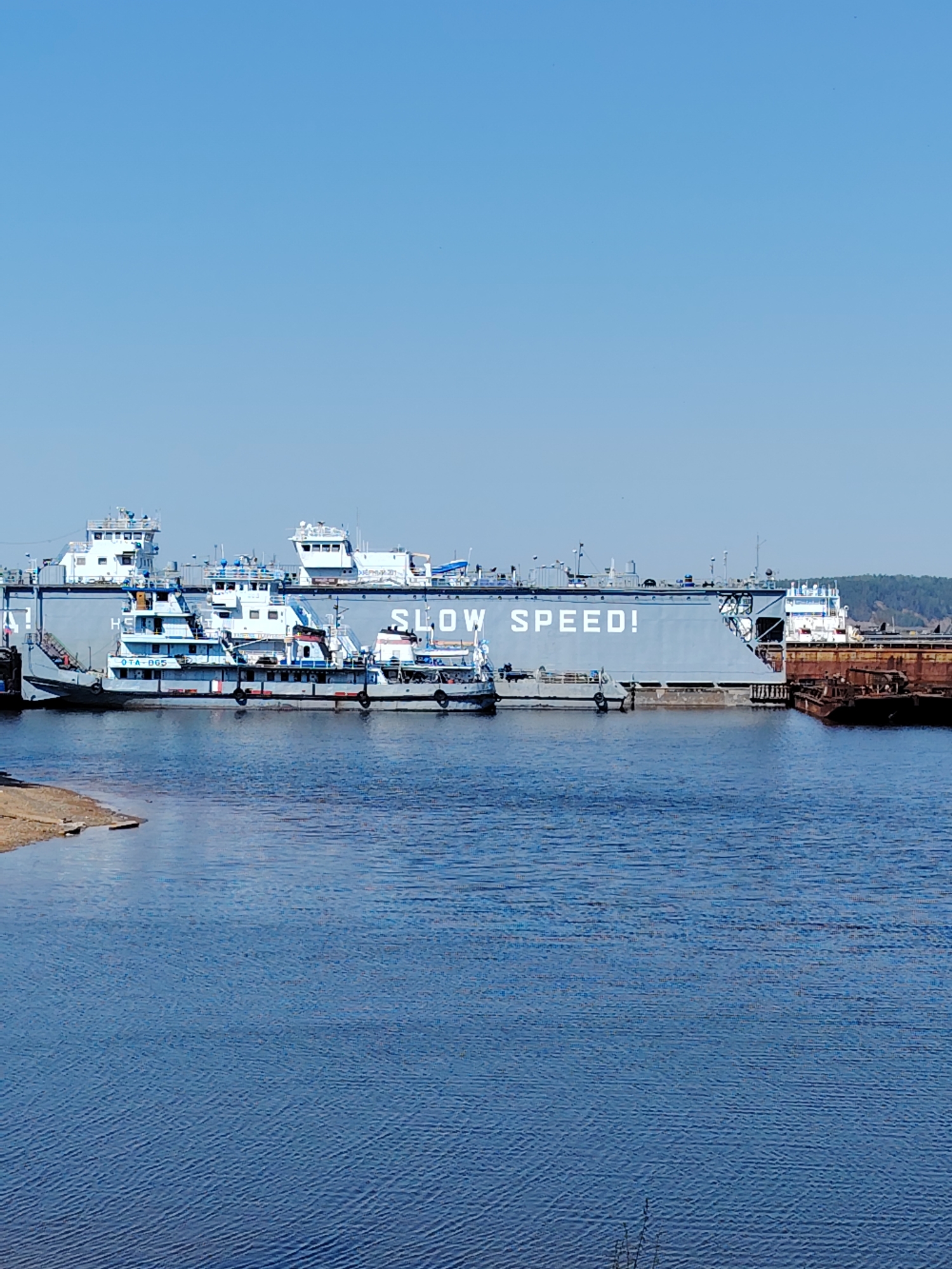 The image shows a river scene in Chaikovsky, Russia. A large, gray barge with the words "SLOW SPEED!" painted in white is docked on the left side of the river. Several smaller tugboats are alongside the barge. The water is a deep blue, and the sky above is clear and light blue. A small patch of sandy shore is visible on the near left. Rust-colored metal structures are docked alongside the barge on its right side. Some trees are visible on the horizon on the far right.