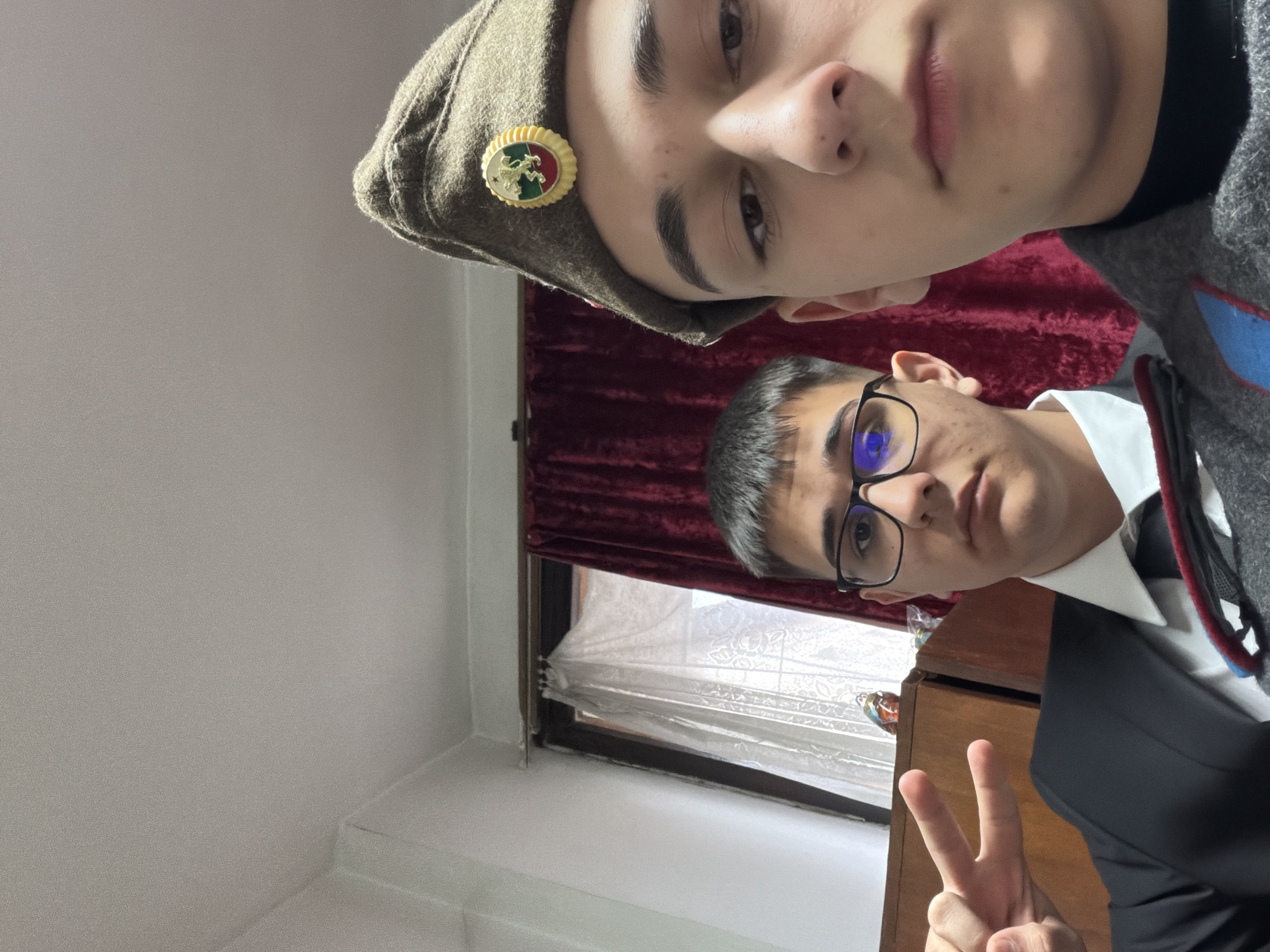 Two young men are posing for a selfie indoors. The person in the upper portion of the frame wears a military-style cap with a badge. The person in the lower portion of the frame wears glasses, a white shirt, and a black jacket. They are giving a peace sign. A window with red curtains is visible in the background. Based on the badge on the military cap, the location is Panagyurishte, Bulgaria.
