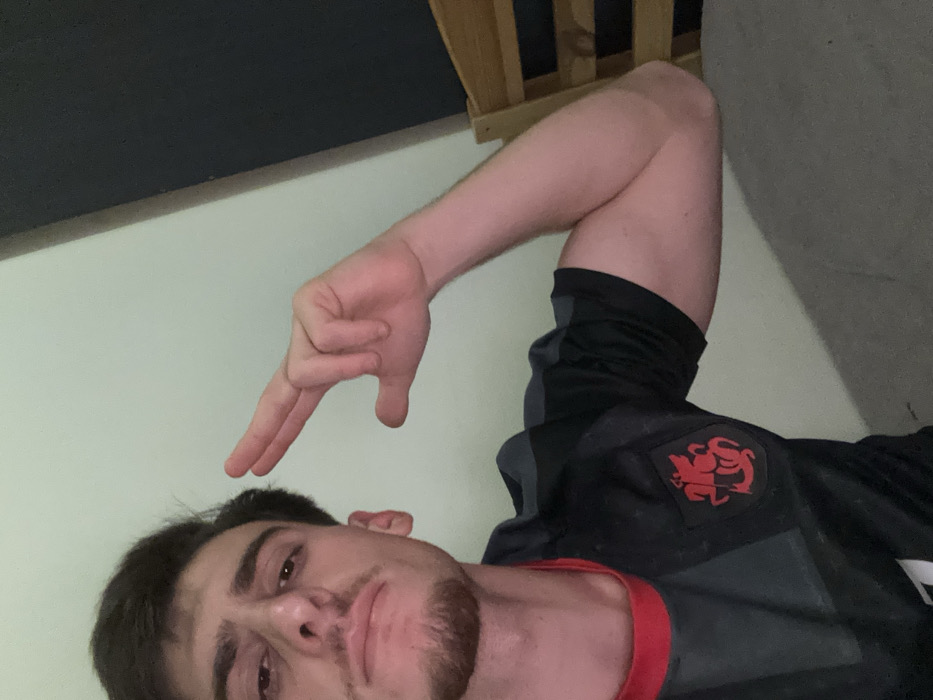A man is positioned in the lower portion of the frame, looking at the camera with a sullen expression. He has short, dark hair and a trimmed beard. Above his head, an arm extends with the hand making a gesture resembling a "loser" sign. The man wears a black and gray athletic shirt with a red trim around the collar. A red lion emblem is visible on the shirt. The background consists of a white wall, a dark upper section of a wall, and a wooden structure.