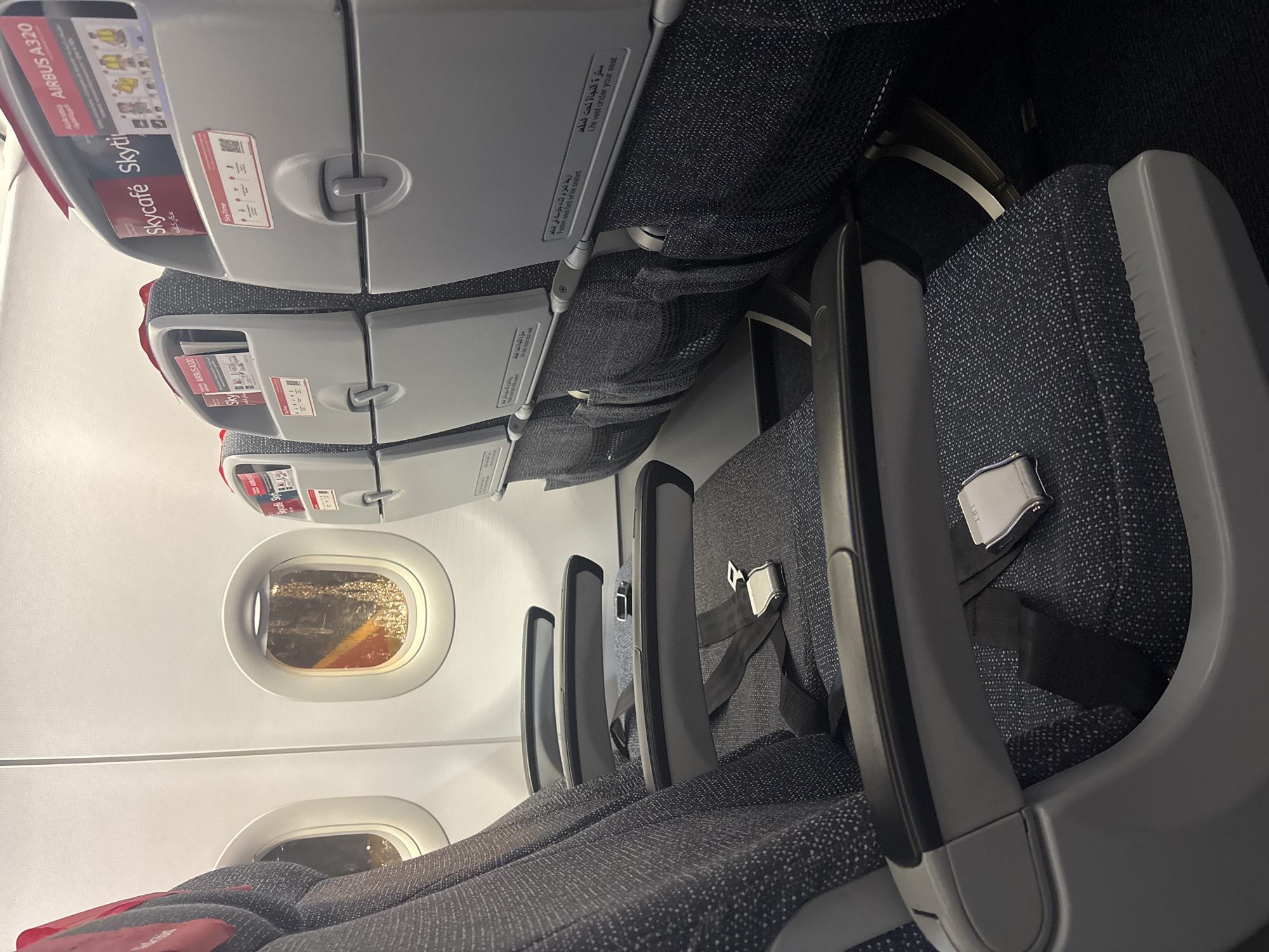 The image displays the interior of an Airbus A320 aircraft cabin, specifically an economy class section, potentially at, departing from, or en route to the city of Katunayake, Sri Lanka. The perspective is from a passenger seat, looking forward and slightly towards the left side of the aisle.
On the left, three rows of passenger seats are visible. The seats are upholstered in a dark blue-grey fabric with a fine white speckled pattern. The seat in the immediate foreground features a visible black seatbelt with a polished silver buckle marked "LIFT." The armrests are constructed from grey plastic with a darker, textured top surface.
The back of the seat directly in front of the viewer is a light grey plastic shell. It includes a closed, foldable tray table and a fabric mesh storage pocket located below it. Above the tray table, a compartment holds several documents: a red magazine titled "Skycafé" in English and Arabic, a "SkyTime" publication, and a safety card explicitly identifying the aircraft as an "AIRBUS A320." Below these, further safety information is printed, including "Fasten seat belt while seated" and "Life vest under your seat," presented in both English and Arabic text. A QR code is also visible on this panel.
Two oval-shaped airplane windows are visible on the left side of the fuselage. The light entering through the rightmost window shows warm orange and yellow hues, suggesting either reflections from the aircraft's wing or fuselage, or the presence of artificial lights from a ground environment during dusk, dawn, or night.
The cabin ceiling is a clean, white paneled surface. The floor is covered with a dark-colored carpet. Interior cabin lighting is active, providing an even illumination throughout the visible area. Partially visible on the right is the back of another seat, consistent in design with those on the left, also containing "Skycafé," "Skytim," and "AIRBUS A320" materials. No individuals are present in the image, and no specific actions are taking place. The scene is static, depicting an unoccupied aircraft interior.
