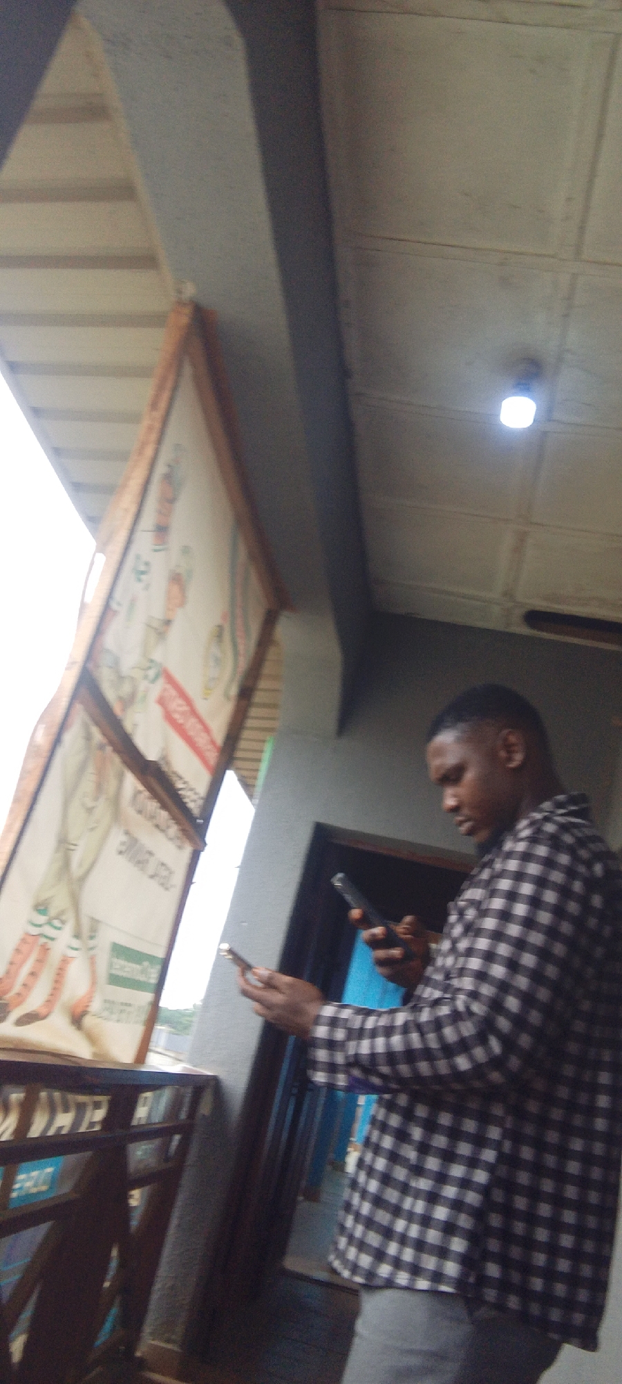 The image shows a young man standing on a balcony, looking at his phone. He is wearing a black and white checkered shirt and grey pants. The man appears to be focused on his phone. In the background, there is a large sign with some drawings and text, and a doorway leading inside. A light fixture is visible on the ceiling. The scene suggests it might be daytime, as the sky is bright. The overall setting appears to be an outdoor area, possibly a marketplace or commercial space, given the sign and the balcony. The man's posture and gaze indicate he is absorbed in his phone usage. There is some text visible on the sign, but it is largely illegible due to the image quality and angle. The man is the primary subject, and his interaction with his phone is the central activity. The location context of Suleja, Nigeria, provides a geographical reference point for the scene.
