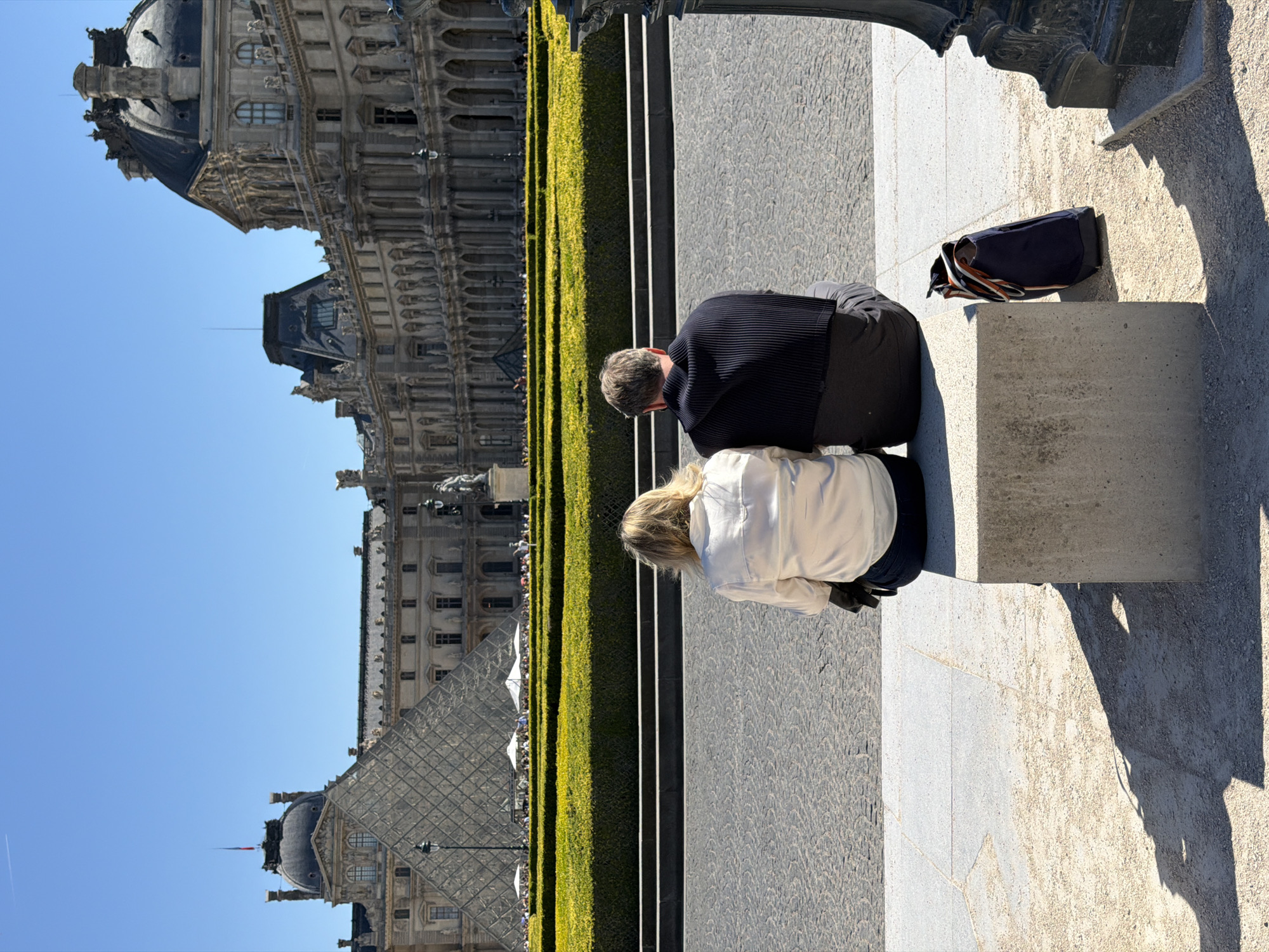 The image shows a man and a woman seated on a low concrete wall in Paris, France. The man is wearing a dark, ribbed top and dark trousers. The woman has blonde hair and is wearing a light-colored jacket. A dark bag sits on the wall next to them. Behind them is a manicured green hedge. In the background is the Louvre Museum, including its glass pyramid and the surrounding buildings. The sky is blue. The image appears to have been rotated 90 degrees counter-clockwise. The surface under the couple is a stone-paved walkway, and the ground to the right of the wall is a light-colored gravel.