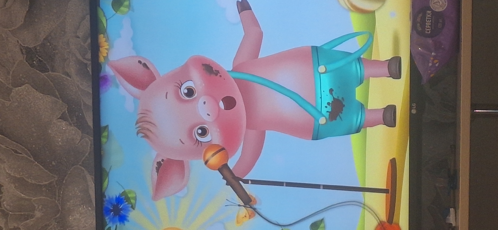 The image depicts a cartoon piglet standing on a yellow surface that resembles sand or dry grass, with a green hill in the background. The piglet is pink, has large ears, and is wearing turquoise shorts with brown mud splatters on them. It has a thin turquoise strap going over its right shoulder and crossing its chest. The piglet is holding a microphone with an orange head, which is attached to a stand. A small, orange butterfly with black markings is perched on the microphone. The piglet has a surprised expression with its mouth open and mud splatters on its cheeks. In the upper left of the image, there is a blue flower and a yellow sun with rays. The background transitions to a light blue sky with white clouds. The image appears to be a screen capture from a digital animation or video game. The bottom of the image shows the bottom bezel of a television screen with the "LG" logo visible, and a purple package with Cyrillic text, likely "СЕРВЕТКИ" (wipes), is placed on a shelf below the screen. There is no identifiable information within the image to suggest a location in Bila Tserkva, Ukraine.