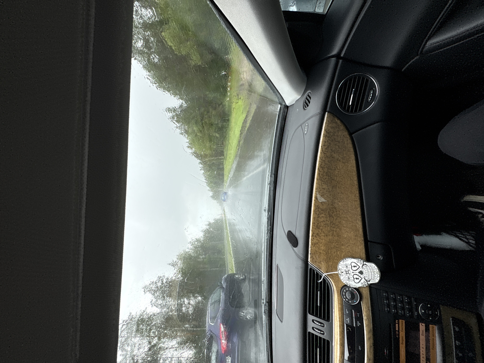 The image is taken from inside a car looking out through the windshield on a rainy day. The windshield is wet with raindrops. Outside, there is a road with cars driving on it. The road is surrounded by trees. The dashboard of the car is visible in the foreground, with a skull-shaped air freshener hanging from the rearview mirror. The location is stated as Гацукоўскі сельскі Савет, Belarus.
