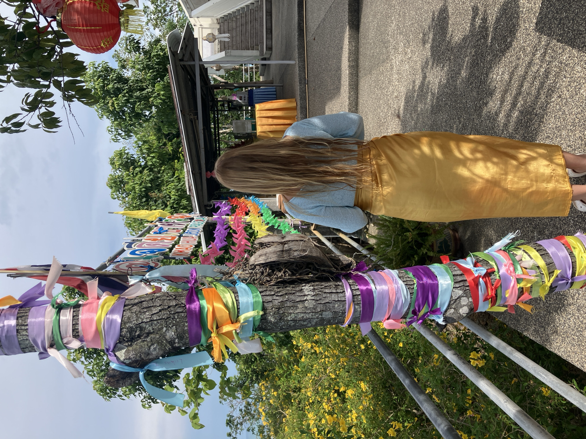 A person with long blonde hair, viewed from behind, stands on a paved pathway in front of a tree adorned with colorful ribbons. The person wears a light blue long-sleeved shirt and a long, shimmering gold skirt. The tree is wrapped with ribbons of various colors, including purple, pink, green, yellow, and blue, tied in bows. To the left of the tree, a metal railing runs along a slope covered in blooming yellow flowers and green foliage. In the background, there is a covered structure with tables, a set of stairs leading upwards, and trees. A red lantern hangs from a tree branch on the right side of the image. A sign with Thai writing is visible behind the tree. The scene appears to be outdoors, possibly in a temple or cultural site, with a glimpse of the sea in the distance on the far left.