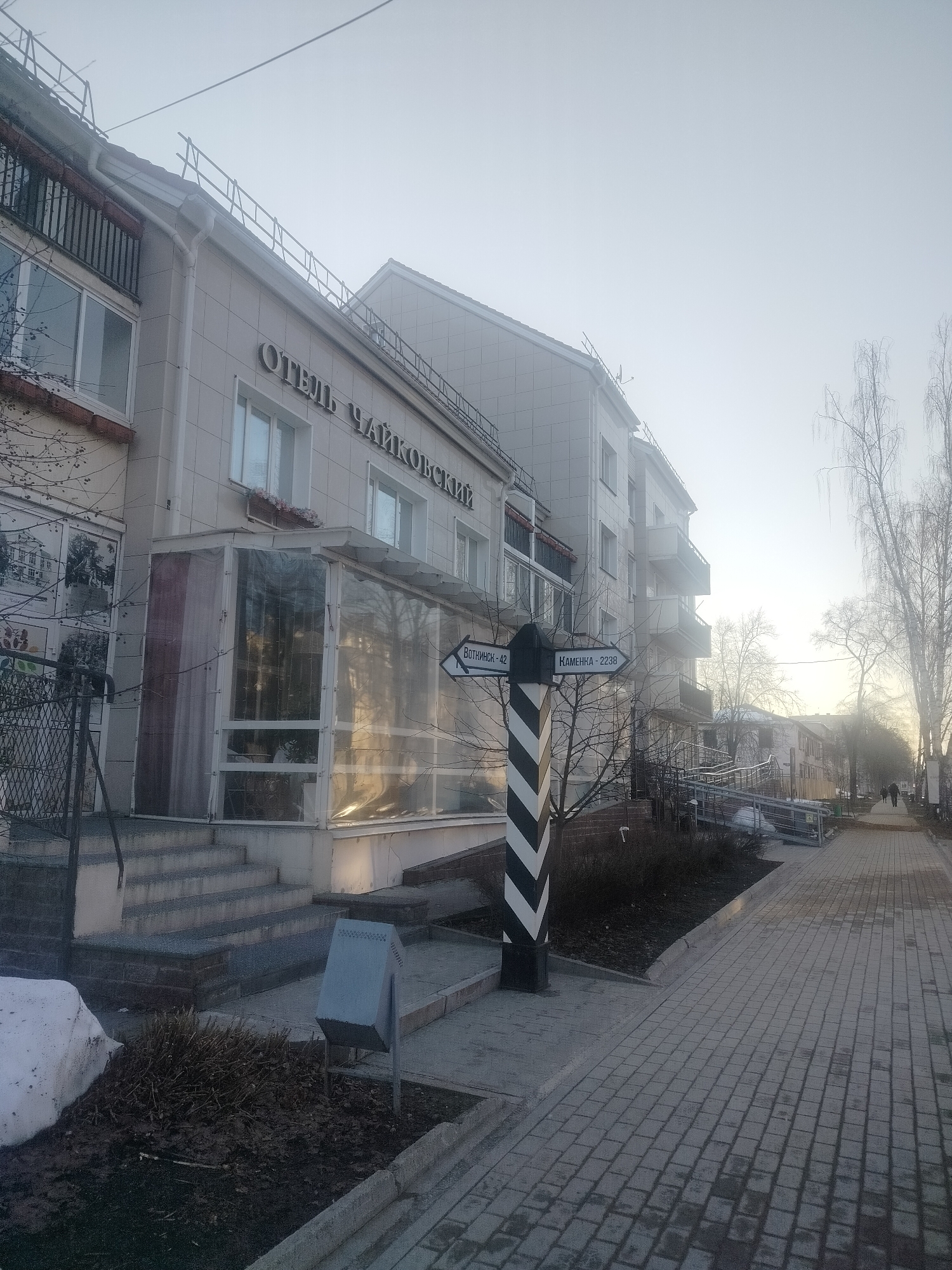 The image depicts an outdoor scene in what appears to be a residential or commercial area of Chaikovsky, Russia. The sky is overcast. The primary focus is on a building that houses the "Отель Чайковский" (Hotel Tchaikovsky). The building is multi-storied with light-colored paneling. It has an enclosed, glassed-in extension at ground level.
A black and white striped signpost stands beside the sidewalk. The signpost has two arrows pointing in opposite directions, labeled "Воткинск - 42" (Votkinsk - 42) and "Каменка - 2238" (Kamenka - 2238).
A paved sidewalk made of interlocking grey bricks is visible in the foreground. A trash receptacle and landscaping beds are present. Bare trees are visible along the sidewalk. Two people are walking along the sidewalk in the distance. Patches of snow linger near the bottom left corner of the image.
