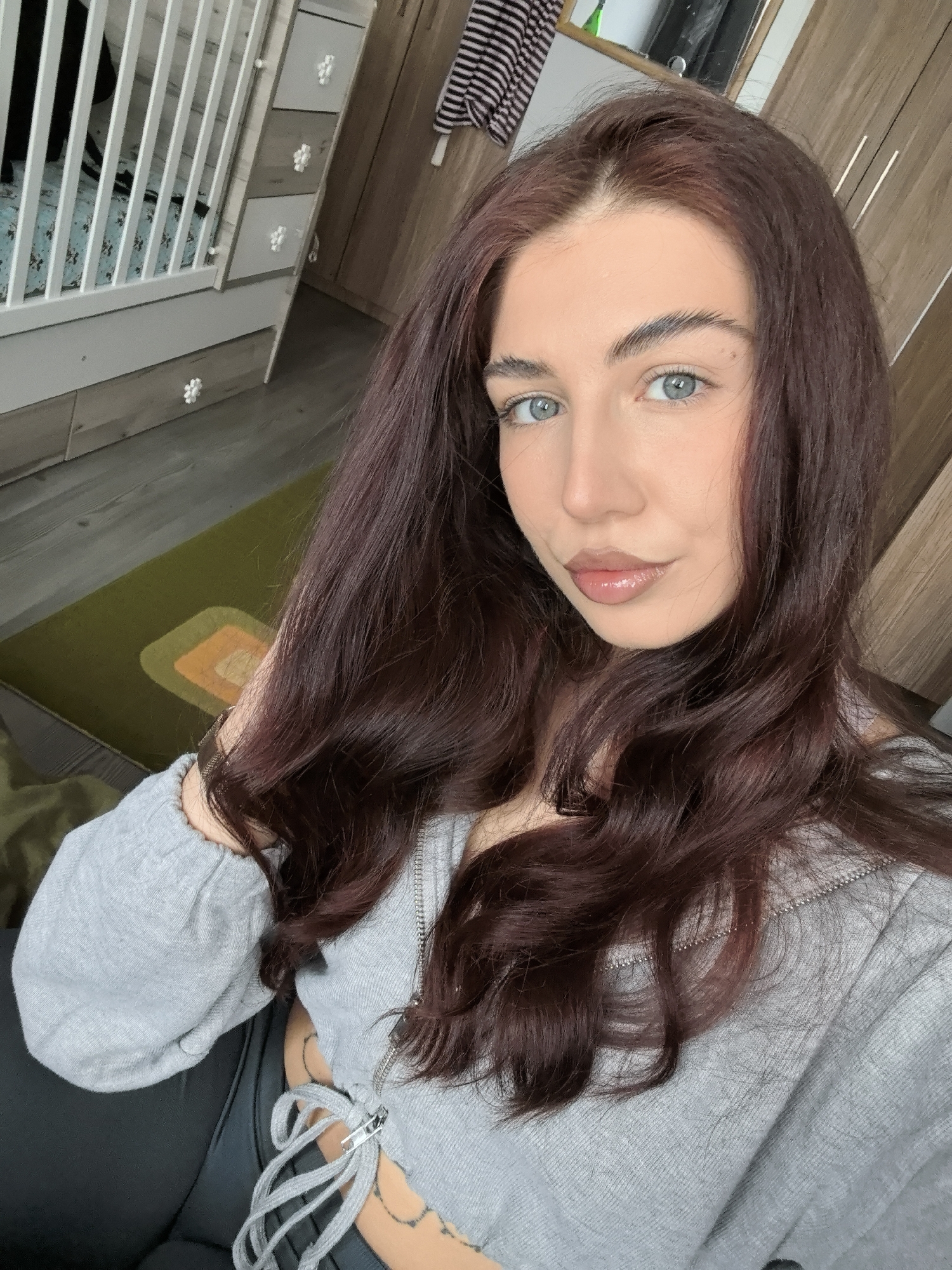 A young woman with auburn hair and light blue eyes is taking a selfie in an interior setting. She is wearing a gray, long-sleeved top and black pants. The top has a drawstring tie at the front. A tattoo is visible on her lower abdomen. Behind her, there's a white baby crib, a wooden dresser, a piece of striped clothing hanging, and a patterned green and yellow rug on a wooden floor. The location is likely within a private residence, but the city of Stara Zagora, Bulgaria, cannot be confirmed from this image alone.
