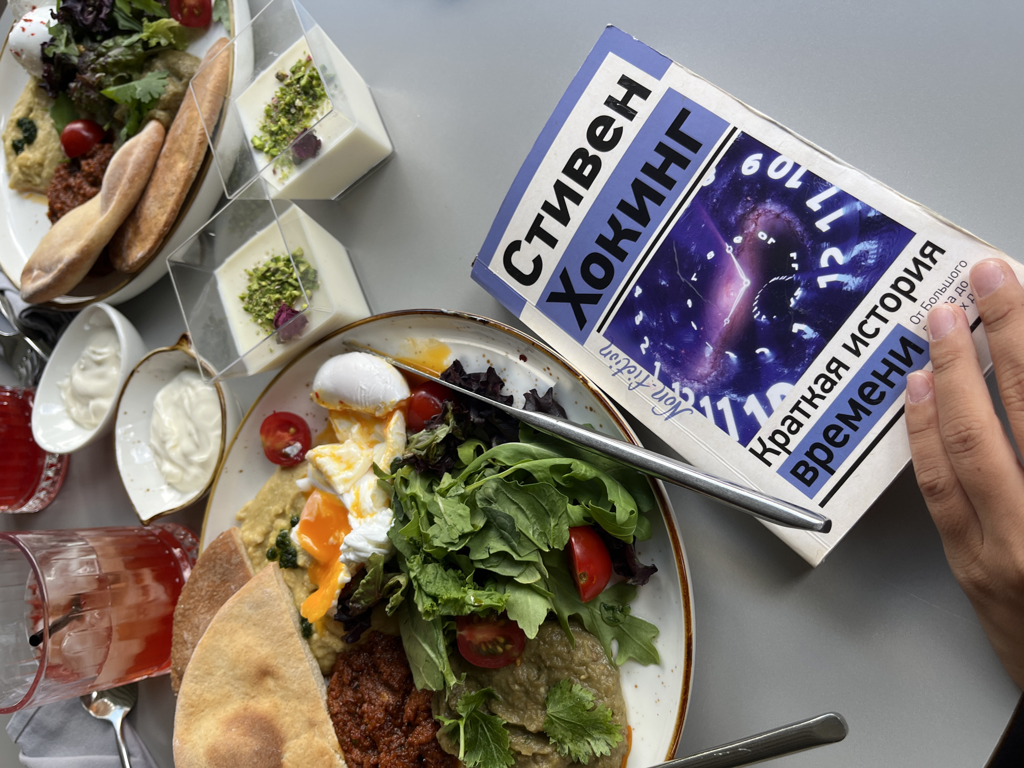 A close-up, overhead shot captures a table setting at a cafe, likely in Almaty, Kazakhstan, with a focus on food and a book. In the foreground, a person's hand rests on a book titled "Краткая история времени" (A Brief History of Time) by Steven Hawking. The book's cover features a celestial image with clock elements.
To the left, a plate of food is visible, consisting of hummus, poached eggs, a reddish spread, and a salad with cherry tomatoes and leafy greens, accompanied by pita bread. To the upper left, a glass of a red beverage with ice and a straw is present. In the background, there are two small clear cups containing a creamy dessert topped with chopped pistachios and a single rose petal. Small bowls of what appears to be yogurt or sour cream are also on the table, along with cutlery. The overall environment suggests a relaxed dining or cafe setting.