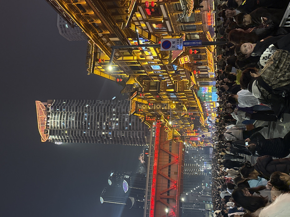 The image presents a dense nocturnal urban scene in the Yuzhong District of Chongqing, China, specifically within what appears to be the Hongyadong Scenic Area. The foreground and midground are occupied by a large, heterogeneous crowd of pedestrians, predominantly adults, standing and moving on a paved street or plaza.
To the right, a series of multi-story buildings constructed in a traditional Chinese architectural style extend along the street. These structures are characterized by dark wooden framework, white wall sections, and ornate, multi-tiered roofs with upturned eaves. They are extensively illuminated with warm yellow and orange lights, highlighting intricate details and numerous suspended red lanterns. Commercial signage, including some with vibrant neon, is visible on these buildings. A blue and white circular sign, bearing a red diagonal line and Chinese text, stands vertically near the traditional structures.
In the background, a modern urban landscape rises. A tall, slender skyscraper with a distinct, curved, red-illuminated architectural feature at its summit is prominent. To its left, additional modern high-rise buildings are visible, some displaying architectural lighting. A substantial, bright red steel bridge with a visible lattice truss structure spans horizontally across the upper-left midground, an elevated component of the city's infrastructure. Streetlights provide localized white illumination across the scene, contributing to the overall bright atmosphere against the dark sky.