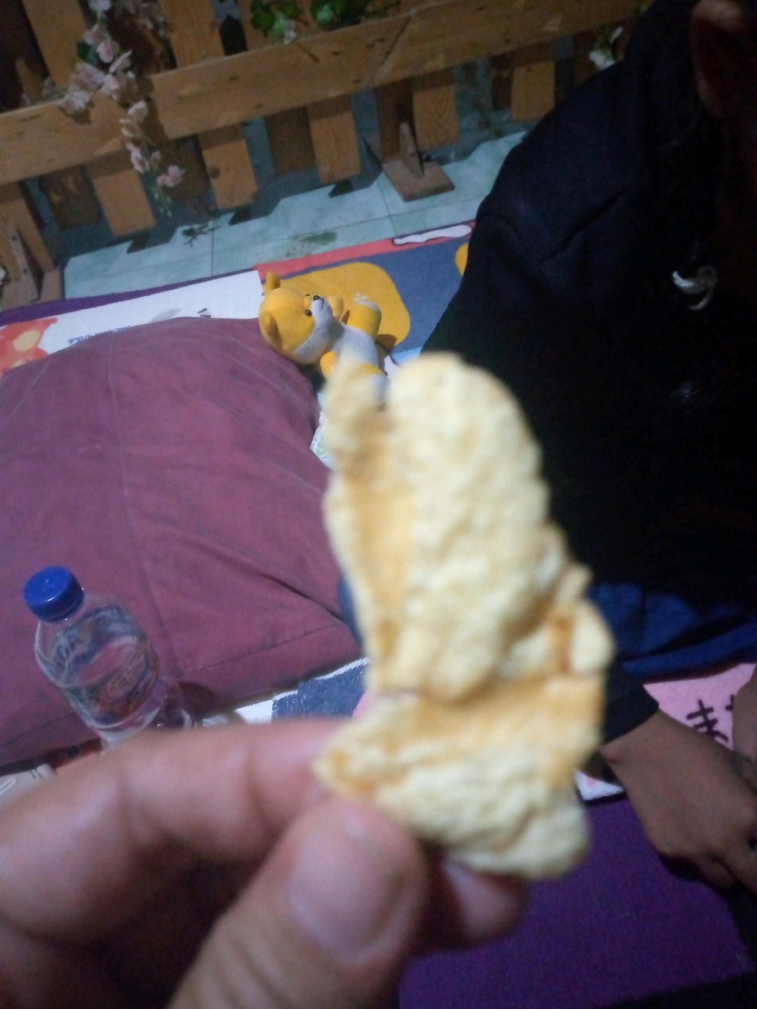 In Sumberwono, Indonesia, a person is holding a piece of puffed snack food in their right hand. The snack is light yellow and appears textured. In the background, a bottle of water with a blue cap is visible on a maroon pillow. Further back, a yellow stuffed toy is positioned on a colorful mat. The environment includes a wooden structure with decorative plants, suggesting an indoor setting or a sheltered outdoor space. The person's left hand is not visible. The image is captured from a close-up perspective, with the snack and the person's hand being the primary focus.