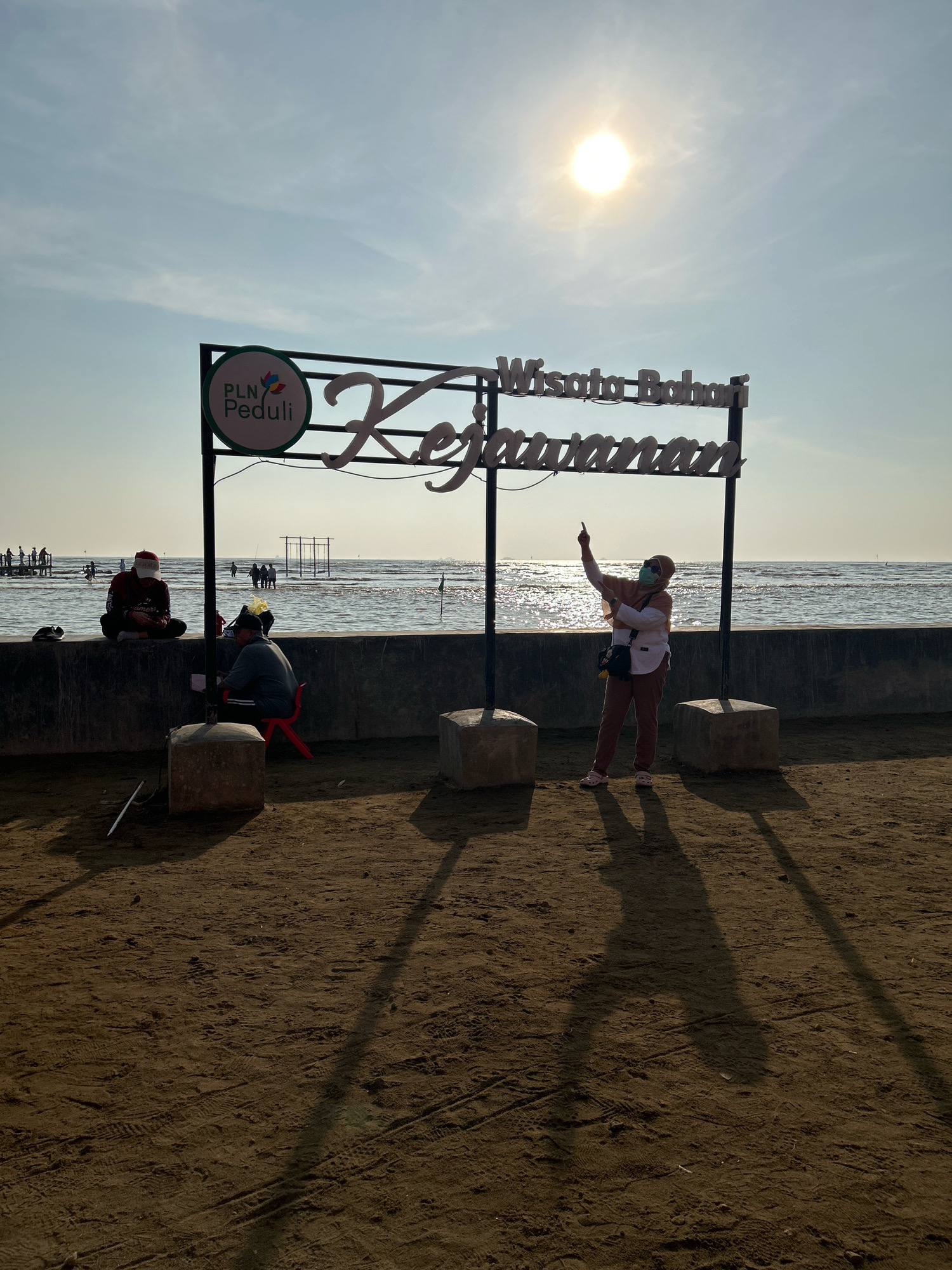 The image shows a sandy beach area with a body of water in the background. A sign with the words "Wisata Bahari" and "Kejamanan" is prominently displayed. Below the sign, there are two men sitting on a concrete wall; one is wearing a red hat and the other a black hat. To the right of the sign, a woman wearing a hijab and mask is posing and pointing. In the distance, there are people walking in the water and structures that appear to be part of a recreational area. The sun is shining brightly in the sky, casting long shadows on the sand. This scene is located at the Kejamanan Beach in Cirebon, West Java, Indonesia.