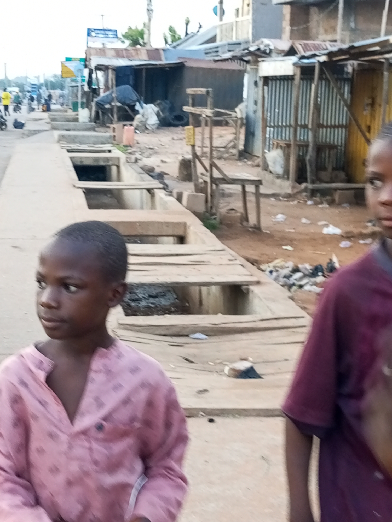 A low-angle, medium shot shows two young boys standing on a street in Tempure, Nigeria. The boy on the left is in the foreground, facing forward and slightly to the left, wearing a pink patterned shirt. His skin is dark, and his hair is short. He has a neutral expression. To his right, the second boy is partially visible, facing the camera and looking off to the right. He is wearing a purple shirt. The background shows a dusty street with open concrete drainage ditches on the left. Along the right side of the street are temporary structures made of corrugated metal and wood, resembling market stalls or informal housing. There is debris, including trash and discarded items, scattered on the ground. In the distance, more buildings and people are visible, suggesting a busy urban environment. The sky is overcast.