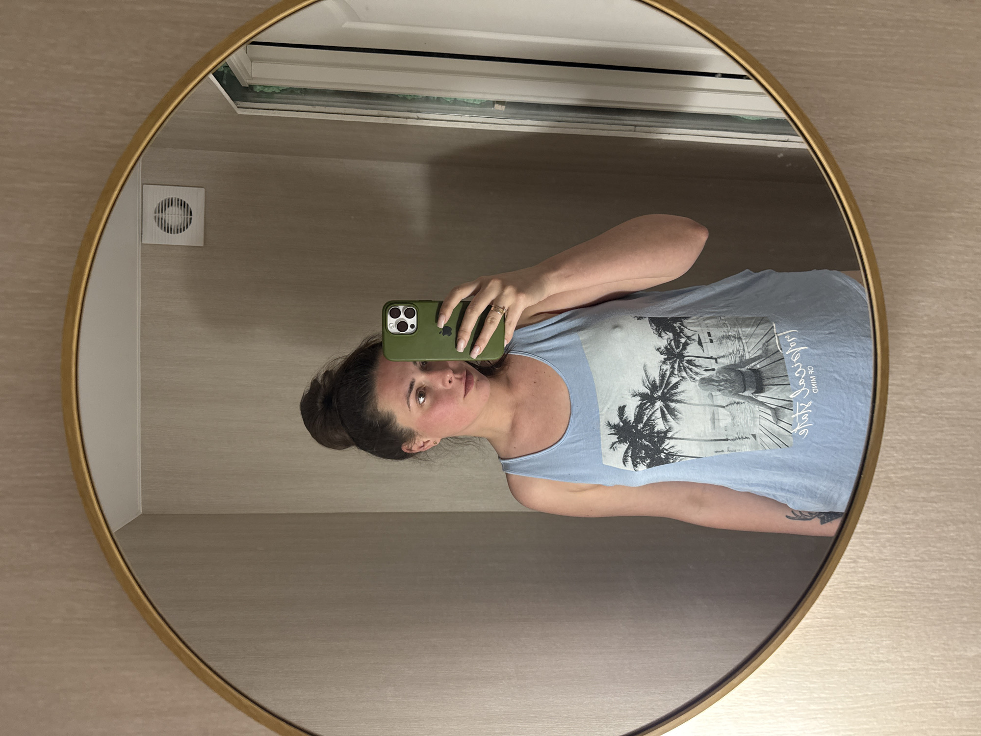 A woman is reflected in a circular mirror with a gold frame. She has her hair pulled up in a messy bun and is wearing a light blue tank top with a graphic of palm trees and a beach scene. She is holding a green smartphone in her right hand, taking a selfie. Her left arm is resting at her side, and a tattoo is visible on her left forearm. The background of the reflection shows a wall with textured wallpaper and a white vent. To the right of the woman, a sliver of a white door or frame is visible. The lighting appears to be artificial.