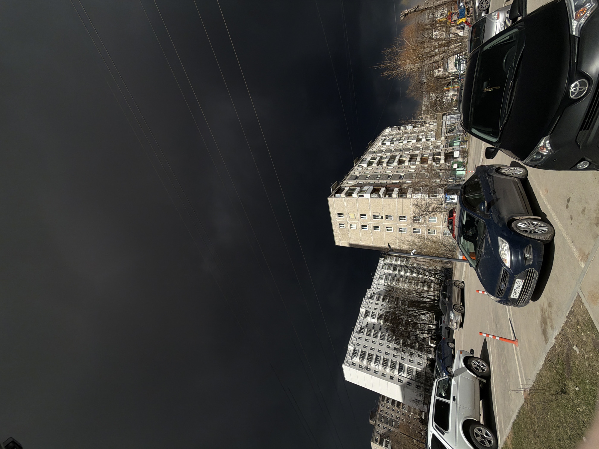 The image captures an urban street scene in Perm, Russia, under a strikingly dark, almost black, sky, suggesting heavy cloud cover or an approaching storm. Numerous utility wires stretch across the upper portion of the frame against this somber backdrop.
On the right, several cars are parked on a paved surface. In the foreground, a dark-colored SUV, possibly a Toyota RAV4, is visible from the front. Behind it, a blue Ford Focus sedan is parked, showing a Russian license plate "T483 BA 59". Further back, a white Lada Niva-like vehicle and other cars are present. Orange and white striped bollards are positioned on the ground between the parked vehicles. Despite the dark sky, some areas of the ground and cars are illuminated by sunlight, suggesting a partial clearing or localized weather event during the day.
To the left and in the background, several multi-story residential apartment buildings, with numerous windows and balconies, line the street. These buildings appear to be characteristic of Soviet-era architecture. Bare trees, without leaves, are interspersed among the buildings and parked cars, indicating the season is likely late autumn, winter, or early spring. No specific activity or people are discernible, but the scene conveys a quiet, dramatic moment in a city environment.