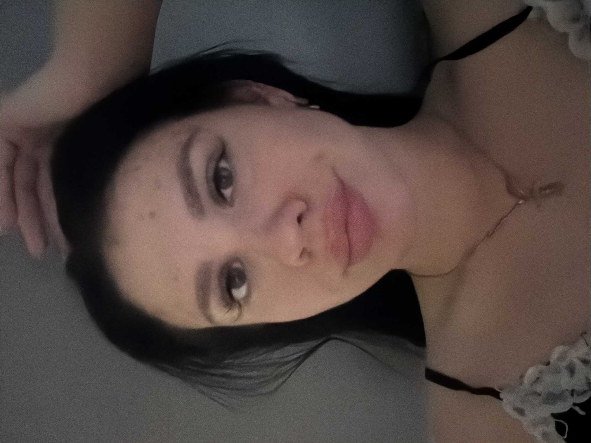 The image is a close-up selfie of a young woman with dark hair, looking directly at the camera. She has full lips and is wearing a necklace. Her right hand is visible resting on her head, and her left arm is bent, with part of a black strap and white lace trim visible on her shoulder. The background appears to be a plain wall. There is no identifiable information within the image that suggests a location in the city of Фоки, Russia, or any specific city.