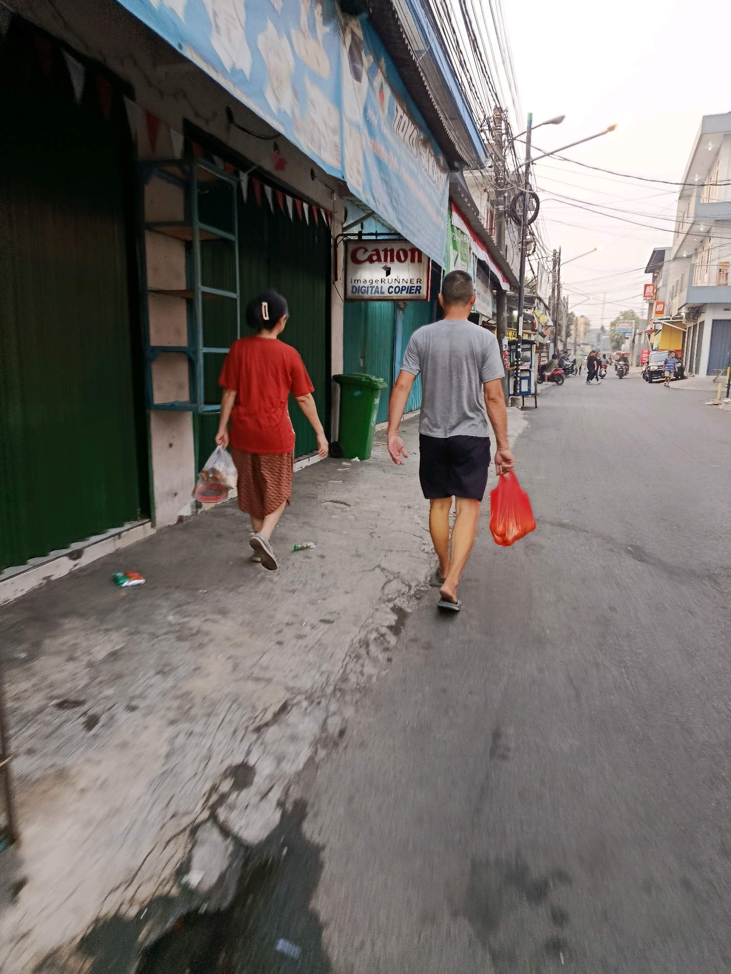 This is a horizontal, slightly low-angle, eye-level shot looking down a street in Bekasi, Indonesia. The perspective suggests the photographer is walking. Two people, a woman in the foreground and a man behind her, walk away from the camera down the right side of a paved street. The woman wears a red t-shirt and a patterned skirt and carries a plastic bag in her right hand. The man wears a grey t-shirt and black shorts and carries a bright red plastic bag in his right hand. Both are walking on the sidewalk to the left of the street. Shops line the left side of the street, their shutters down and decorated with flags. A green trash bin is visible on the sidewalk near the woman. The street is mostly empty, with some distant cars and motorbikes. The sky is overcast and bright.