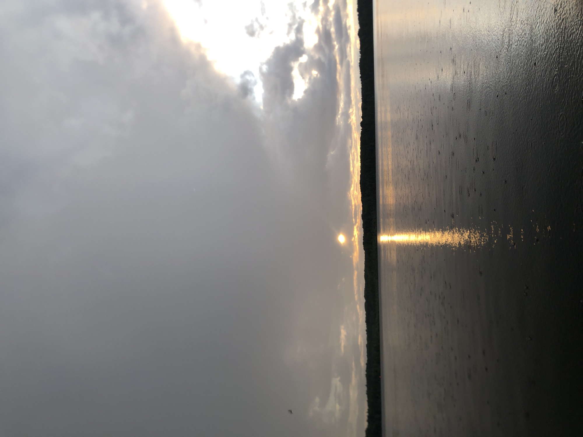 This media file depicts a serene natural scene, likely at dusk or dawn, given the low angle of the sun. The sun is partially obscured by clouds, casting a golden, diffused light across the sky and reflecting shimmeringly on the water. The water appears to be a large body, possibly a lake or a wide river, with a dark, tree-lined shore visible in the distance as a thin, dark strip. The surface of the water shows ripples and small circles, suggesting that it is raining lightly. The sky is a mix of grey clouds and patches of brighter, sunlit areas. The overall mood is tranquil and contemplative, evoking a sense of calm and natural beauty. There is no visible text in the image.