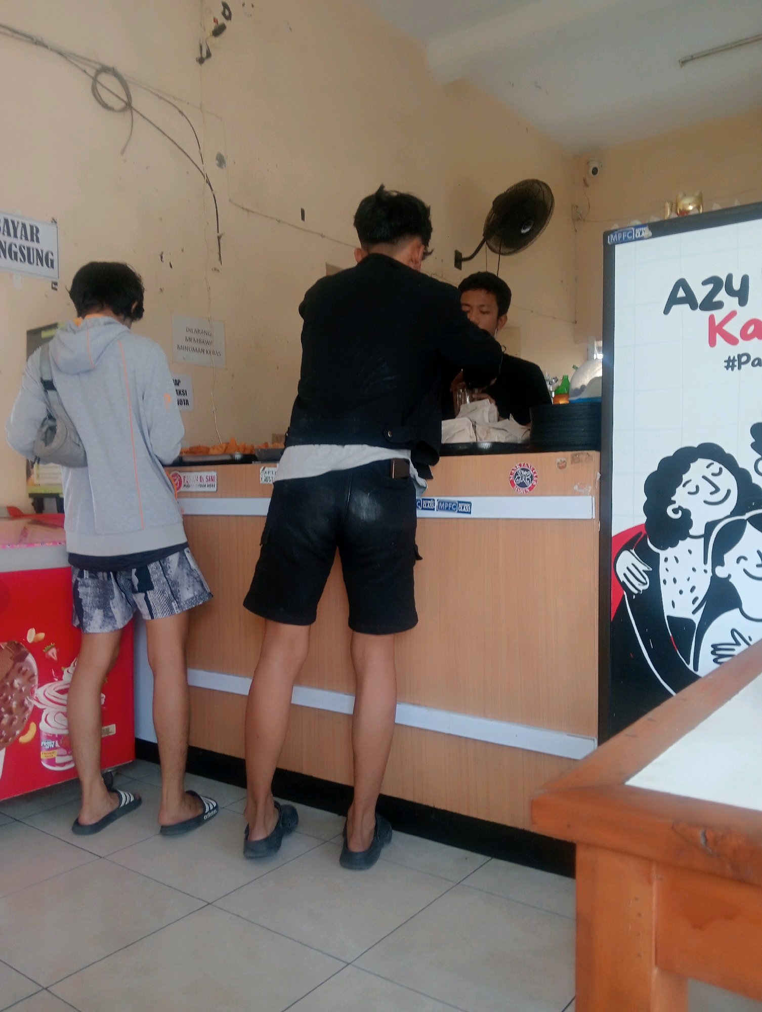 The image depicts an indoor scene at a food stall or small eatery in Plosojenar, Indonesia. Two young men are standing at the counter, with one appearing to be a customer and the other potentially an employee or another customer. The customer, on the left, is wearing a grey hooded jacket over a black t-shirt and patterned shorts, with a grey bag slung over his shoulder. He is facing away from the camera. The second young man, in the center, is wearing a black jacket over a white t-shirt and black shorts, also facing away from the camera. He is leaning over the counter, possibly interacting with someone behind it, though only a glimpse of another individual is visible. The counter is made of light-colored wood, with various food items, possibly snacks, displayed on trays. A large red refrigeration unit with ice cream branding is visible to the left of the counter. On the wall behind the counter, there are several signs. One sign, partially obscured, appears to be a price list or menu. Another sign, in Indonesian, reads "DILARANG MEMBAWA MINUMAN KERAS," which translates to "NO ALCOHOLIC DRINKS ALLOWED." A wall-mounted fan is visible on the upper right side of the wall. To the right of the counter, a large whiteboard or poster features text and illustrations, including the words "A24" and "Ka." The floor is tiled with square beige tiles. An orange wooden table is partially visible in the lower right foreground. The overall environment suggests a casual, local establishment.
