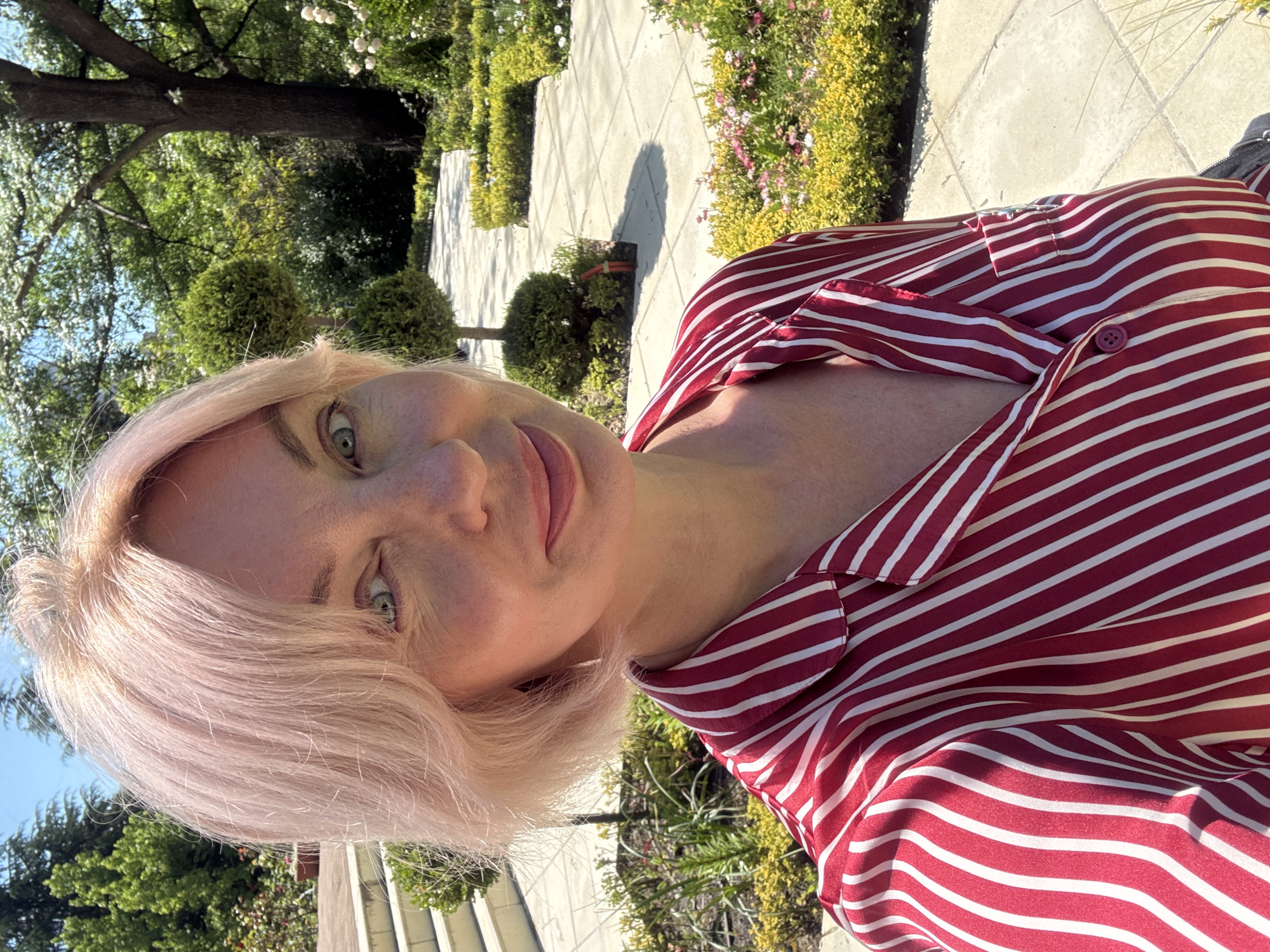 A woman is captured in a sunlit outdoor selfie, her head tilted, looking directly at the viewer with a soft smile. She has light-colored, possibly pale blonde or pink-tinted hair, and light-colored eyes. She is wearing a button-down shirt with horizontal red and white stripes.
The background shows a verdant garden or park setting under bright daylight. A light-colored paved walkway or patio with square tiles extends behind her, bordered by manicured green bushes, some trimmed into spherical topiary shapes. Further back, lush green trees with dense foliage are visible, suggesting a rich natural environment. The strong sunlight creates clear, defined shadows on the paved ground, indicating a bright and clear day. No specific activities are taking place other than the woman posing for the photo. The overall mood is peaceful and pleasant. There is no visible text in the image. The setting aligns with a beautiful garden or park typical of Sochi, Russia.