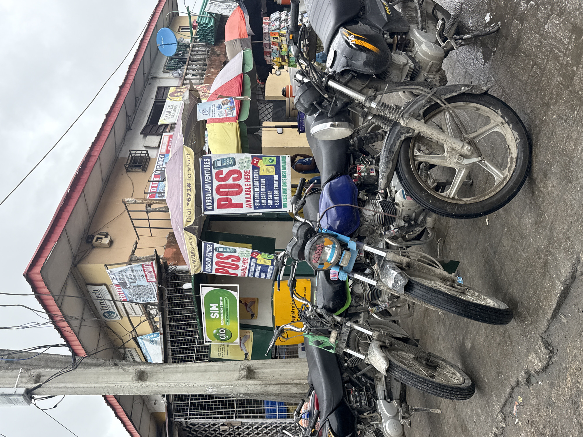 A street-level, medium shot captures a row of several motorcycles parked on a wet, muddy, asphalt surface in Ado, Nigeria. The motorcycles, mostly of the "Okada" style, are heavily laden with dust and mud, suggesting recent rain or an unpaved road. In the background, a building with a yellow facade features several signs, including "Ilbrotech" and "SIM Registration Center" from "Glo." Another prominent sign advertises "Warsalam Ventures POS" services. Umbrellas are set up in front of the building, possibly to shelter vendors or customers. The scene appears to be a busy commercial area, with the motorcycles likely used for transportation and the POS services indicating a hub for financial transactions. The sky is overcast, contributing to the muted lighting of the scene. No people are clearly visible in the immediate foreground or engaged in specific actions.