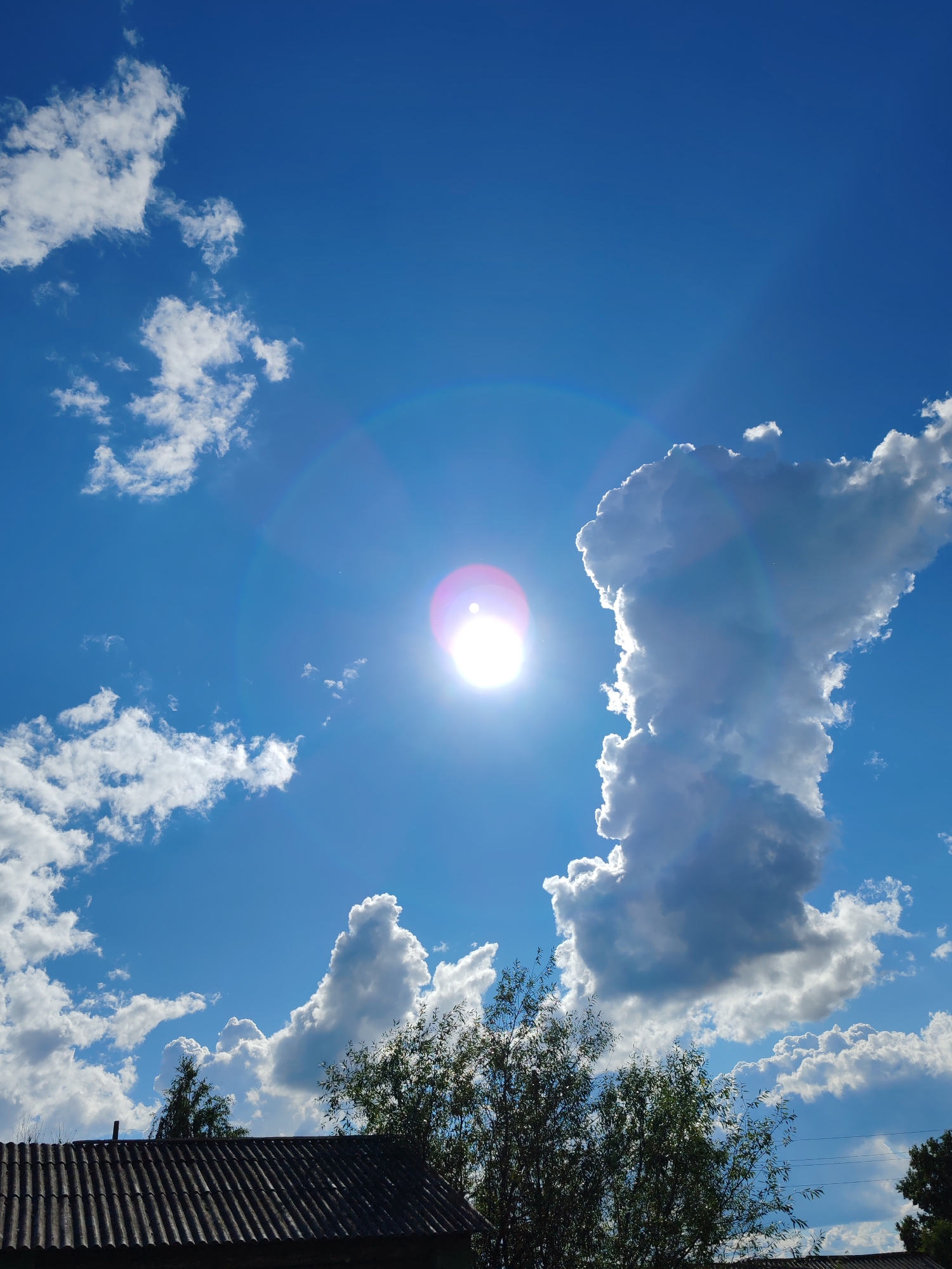 The image shows a bright, sunny day with a clear blue sky filled with fluffy white clouds. The sun is shining intensely, creating a lens flare with a pinkish-purple hue. The clouds are varied in size and shape, with some appearing very dense and illuminated by the sun, while others are smaller and more wispy. The bottom portion of the image features the roof of a building, likely a house, made of corrugated material. Some trees and foliage are visible above the roofline, with branches and leaves silhouetted against the bright sky. The scene appears to be outdoors, possibly in a rural or suburban area. There are no people or visible actions in the image. The overall impression is one of a pleasant summer day.