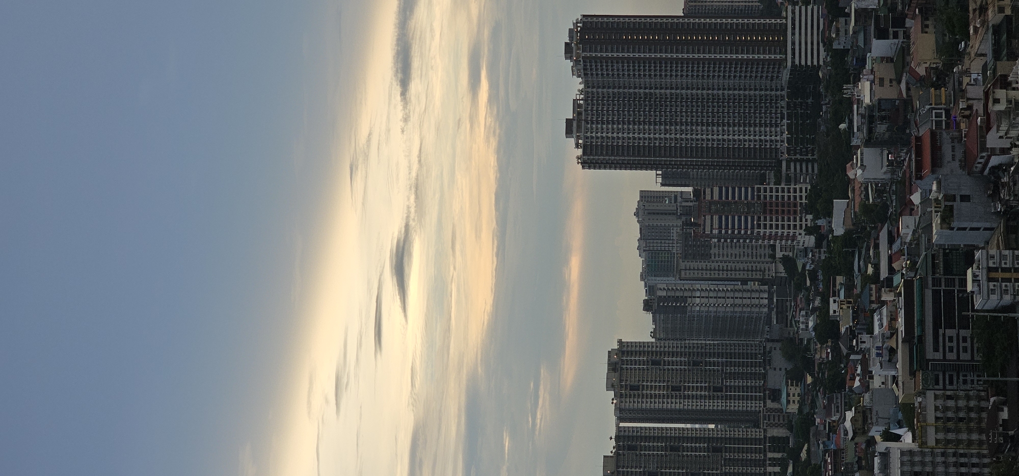 The image captures a cityscape in Makati, Philippines, likely during either sunrise or sunset given the soft, diffused light and the warm hues in the sky. The foreground is dominated by a dense cluster of residential buildings and apartment complexes, varying in height and architectural style. Many of these buildings appear to be high-rise structures, with numerous windows and balconies indicative of urban living.
In the mid-ground, a mix of lower-rise buildings with pitched roofs and modern facades suggests a blend of older and newer developments. Greenery is interspersed throughout, with trees visible between buildings, indicating some residential areas have pockets of vegetation.
The background features a striking array of modern high-rise buildings, characteristic of a central business district. These skyscrapers are tall and rectangular, with a repeating pattern of windows. Some appear to be under construction, with exposed frameworks visible.
The sky is a significant element, transitioning from a soft blue at the top to warmer tones of yellow and orange closer to the horizon, diffused by clouds. The clouds are scattered and appear to be illuminated by the sun, creating a serene and atmospheric effect.
There are no visible people or discernible actions taking place in the image, focusing solely on the built environment and the sky. The scene conveys a sense of a bustling, developing urban area, observed from a vantage point that overlooks a significant portion of the city. The overall impression is one of a densely populated metropolitan landscape under a tranquil sky.