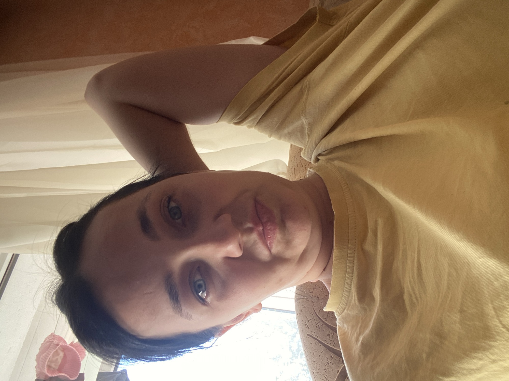 A woman is captured in a close-up, frontal shot, her head tilted back with her left arm behind her head. She wears a yellow t-shirt. Her dark hair is pulled back, and her expression is neutral. The background consists of sheer white curtains and a textured orange wall. Sunlight streams in from a window to her left. The setting appears to be indoors, likely a residential area, with a patterned armchair visible beneath her.