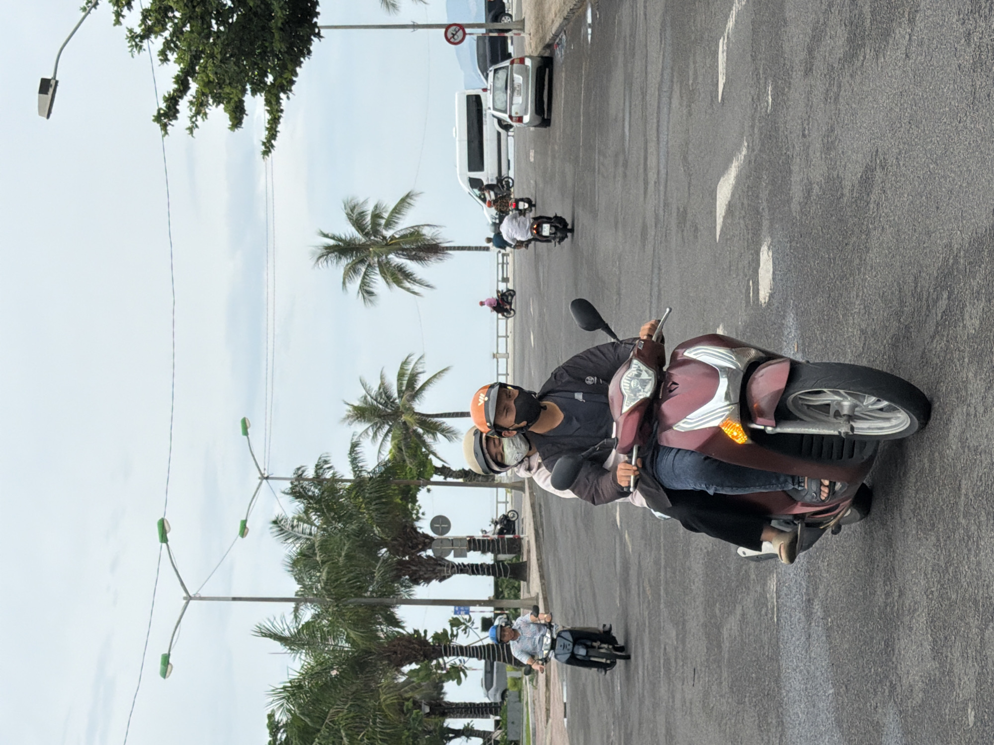 The image shows a street scene in Nha Trang, Vietnam, with several motorbikes and cars on a road. In the foreground, two people are riding on a burgundy scooter. The driver is wearing a black helmet and a black face mask. The passenger behind him is wearing a light-colored helmet and a white face mask. The scooter is moving forward on the asphalt road. In the background, there are palm trees, streetlights, a white van, and other vehicles. The sky is overcast.