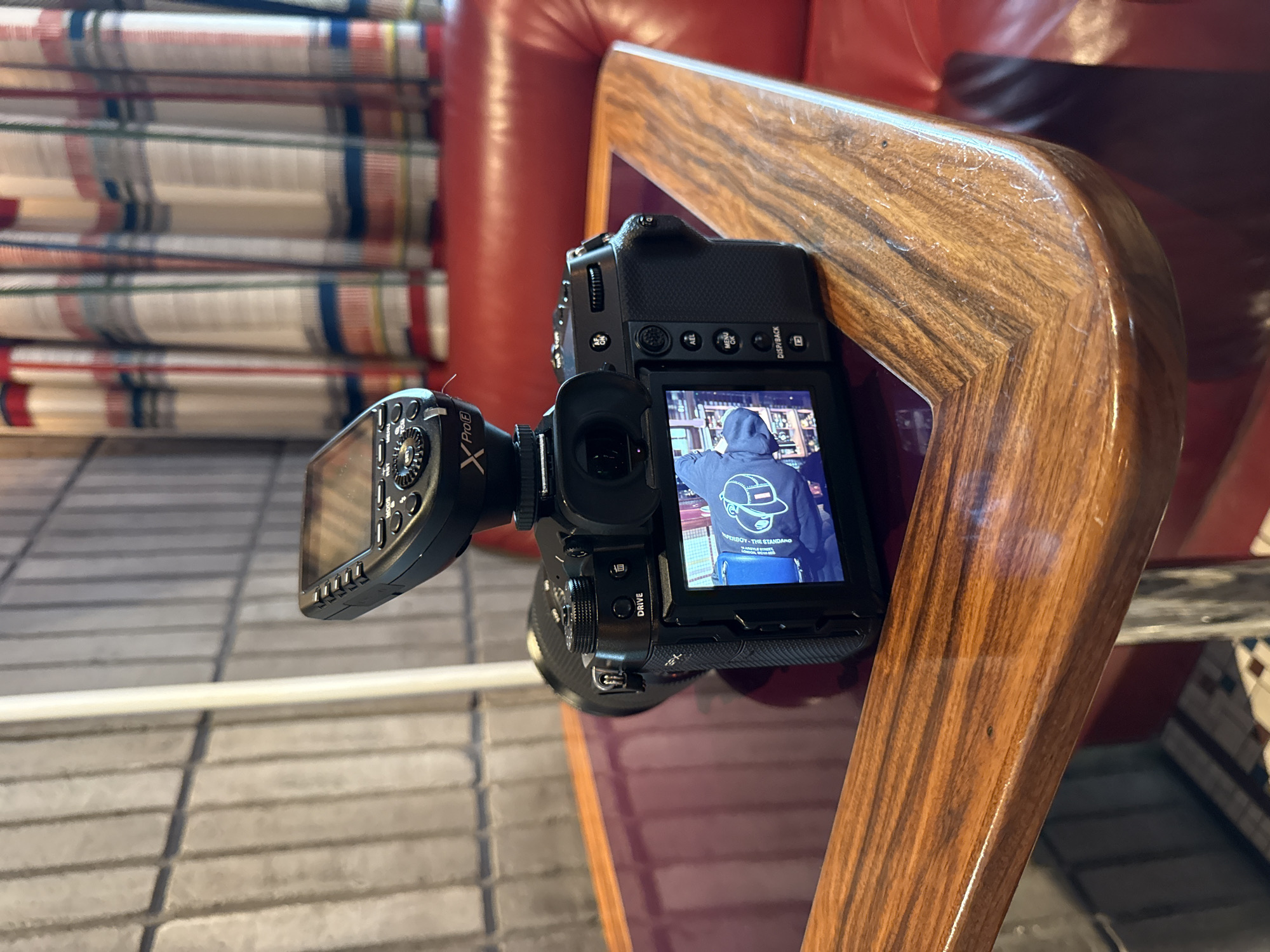 A Fujifilm X-T series mirrorless camera, likely an X-T5, is positioned vertically, resting on a polished wooden armrest or frame. An external Godox XProF flash trigger is mounted on its hot shoe, extending to the left. The camera's rear screen displays a live view or captured image of a person seen from behind, wearing a blue hooded sweatshirt with a distinctive white logo. The logo depicts a cartoon-style figure wearing a cap and includes visible text, appearing to say "SUPERBOY - THE STANDARD" followed by smaller lines of text underneath, possibly "OUR OWN DESIGN" and "PREMIUM QUALITY". The background on the camera's screen shows what appears to be a well-stocked bar or shelves with bottles.
The camera's controls are visible, including "AEL," "MENU/OK," "DISP/BACK," and "DRIVE" buttons on the rear, and dials for shutter speed and exposure compensation on the top. The Godox flash trigger clearly shows "XProF" branding and various function buttons like "GROUP," "MODE," and "ZOOM."
The immediate background of the overall image features a plush, red leather sofa or chair, against which the wooden frame rests. Further in the background, a stack of neatly folded or rolled fabric, possibly blankets or curtains, displays a plaid pattern with white, red, blue, and green stripes. The floor beneath the camera is a light-colored tile or patterned surface. The setting suggests an indoor environment, possibly a hotel lounge, a trendy bar, or a comfortable waiting area in London, where the camera is set up for a photographic session in low light conditions. The camera's position suggests it might be ready to capture a scene or subject within this establishment.