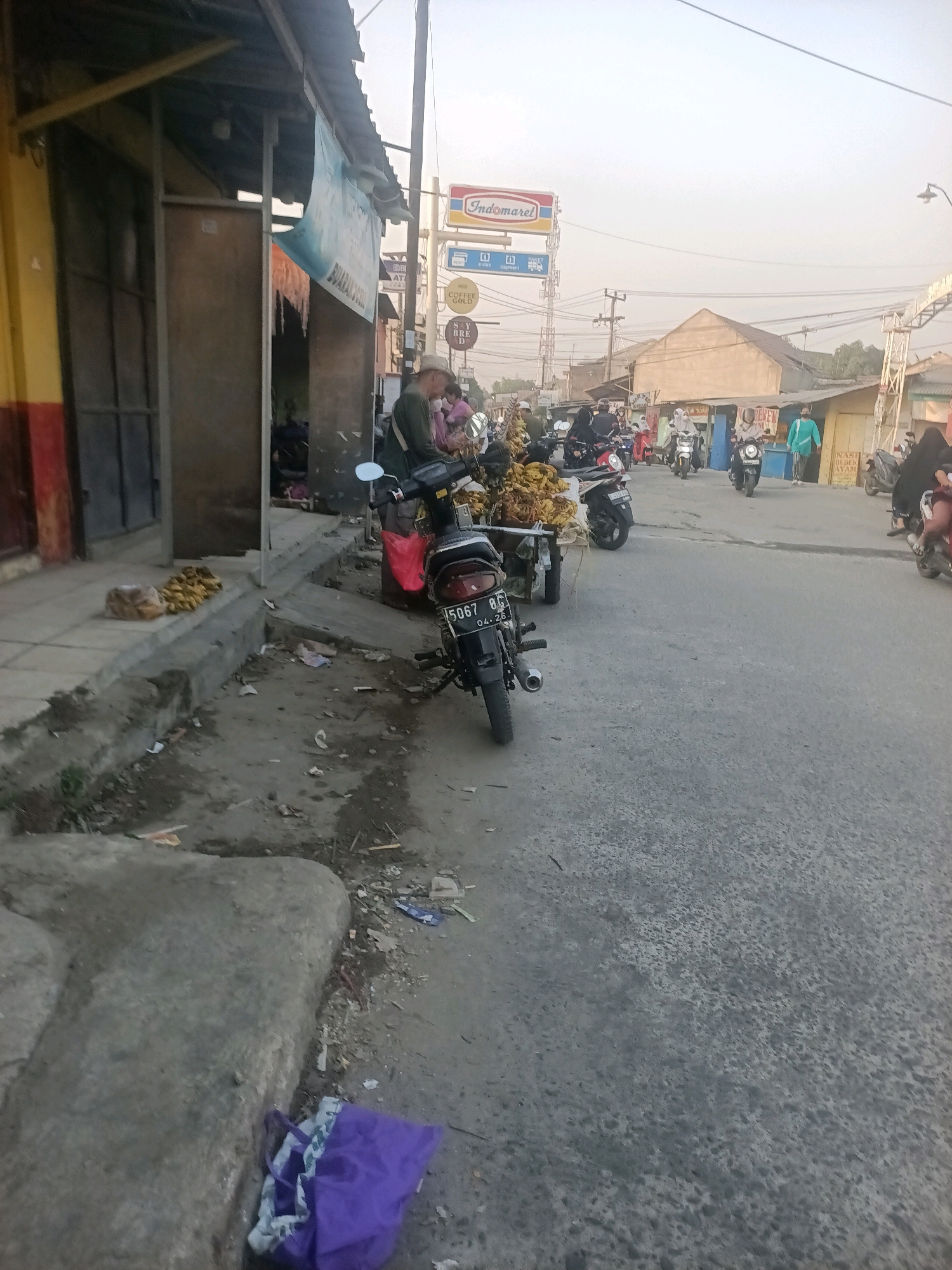 The image depicts a street scene in Wanajaya, Indonesia, with a focus on a commercial area and the surrounding environment. In the foreground, a motorcycle with license plate "5067 BG 04.26" is parked on the right side of the road. To the left, a concrete sidewalk and curb are visible, with a pile of bananas and a few discarded items on the ground. Beyond the sidewalk is a building with a yellow and red facade, featuring an open entrance and a wooden structure.
Further down the street, several other motorcycles are present, some with riders. A person wearing a green top and a white hat is standing next to a motorcycle with a red bag attached. In the background, a street vendor is seen with a cart laden with bananas. Various shops line the street, including one with an "Indomaret" sign, a convenience store chain. The street is paved and appears to be a busy area with pedestrian and vehicular traffic. The overall atmosphere suggests a typical urban market or commercial district. The sky is overcast, and the lighting indicates it is likely morning or late afternoon.