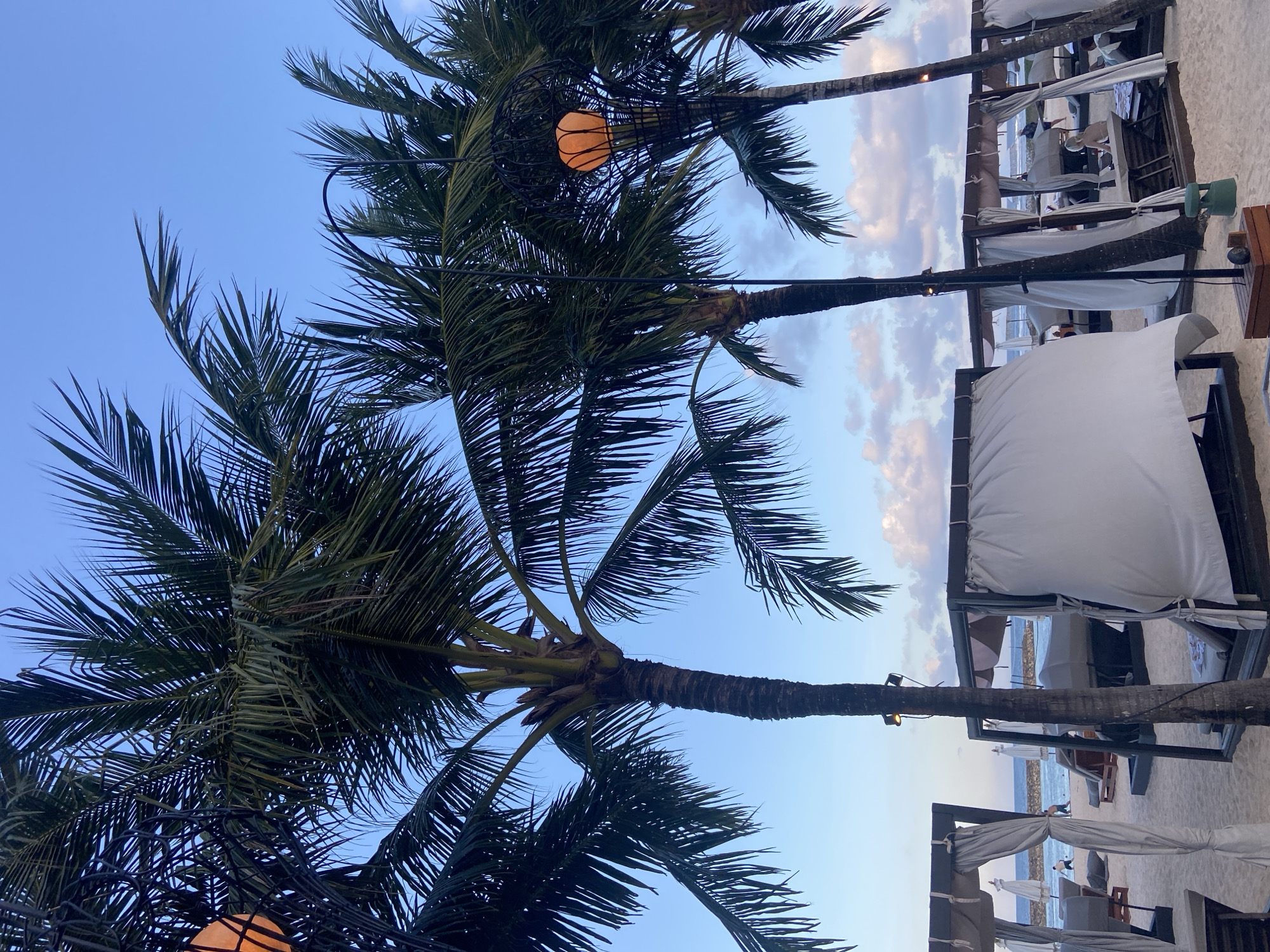The image shows a beach scene in Kuta Selatan, Indonesia. Tall palm trees dominate the foreground, with string lights hanging from their fronds. In the background, cabanas with white curtains line the beach. The sky is a mix of blue and light clouds. The scene is oriented vertically. The beach is sandy and there are chairs and umbrellas visible near the water.