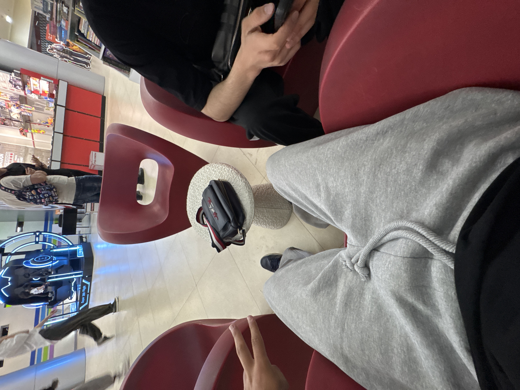 The image captures a first-person perspective from someone seated in a modern red chair within an indoor commercial or entertainment setting in Yerevan, Armenia. In the immediate foreground, the photographer's hand forms a peace sign, and their leg, clad in grey sweatpants with a visible drawstring, is stretched out.
Opposite the photographer, another person is seated in an identical red chair, wearing a black top and pants, engrossed in a smartphone held in their hands. Between these two individuals is a small, round, white textured table, upon which a black shoulder bag with red and white stripe details is placed.
The background shows a lively scene. To the left, a person wearing a white top and dark pants, carrying a patterned shoulder bag, walks past red retail shelving displaying various items, possibly books or merchandise. A red box with the text "POLYGONE" is also visible in this area. Further into the background, a large, blue-lit arcade machine stands out. The machine clearly displays the words "SUPER 360 FLIGHT," indicating a virtual reality or simulation game. Another person, appearing somewhat blurred due to motion, is visible near this arcade machine. The floor is light-colored and tiled. The overall environment suggests a casual hangout or waiting area within a shopping mall or leisure center, with artificial lighting indicating an indoor setting during the day or evening.